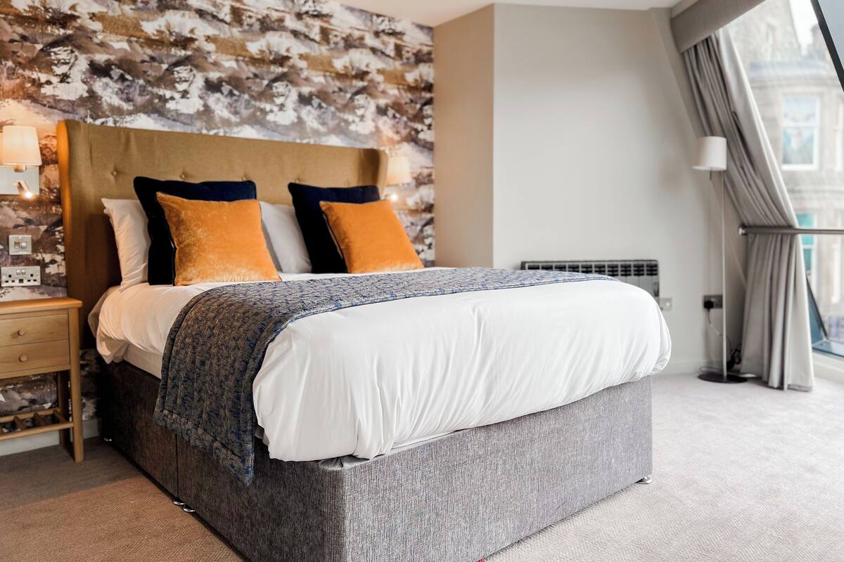 Ein modernes Hotelzimmer mit einem großen Bett, einer Bettseite mit strukturierter Gestaltung und Blick auf die Stadt durch große Fenster.