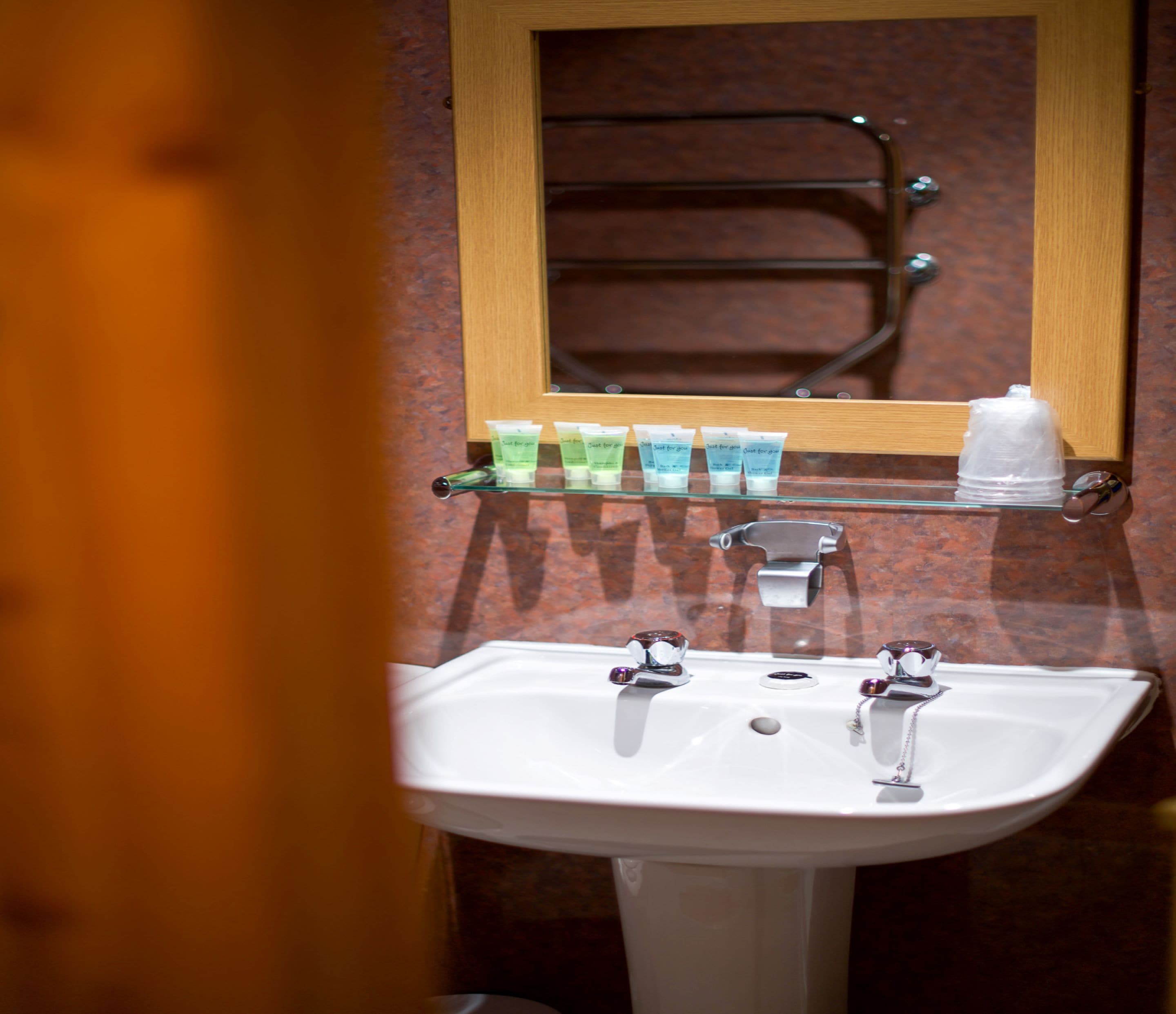 A bathroom with a white sink, two faucets, and a mirror above it. There are four green and blue tubes on a shelf above the sink.