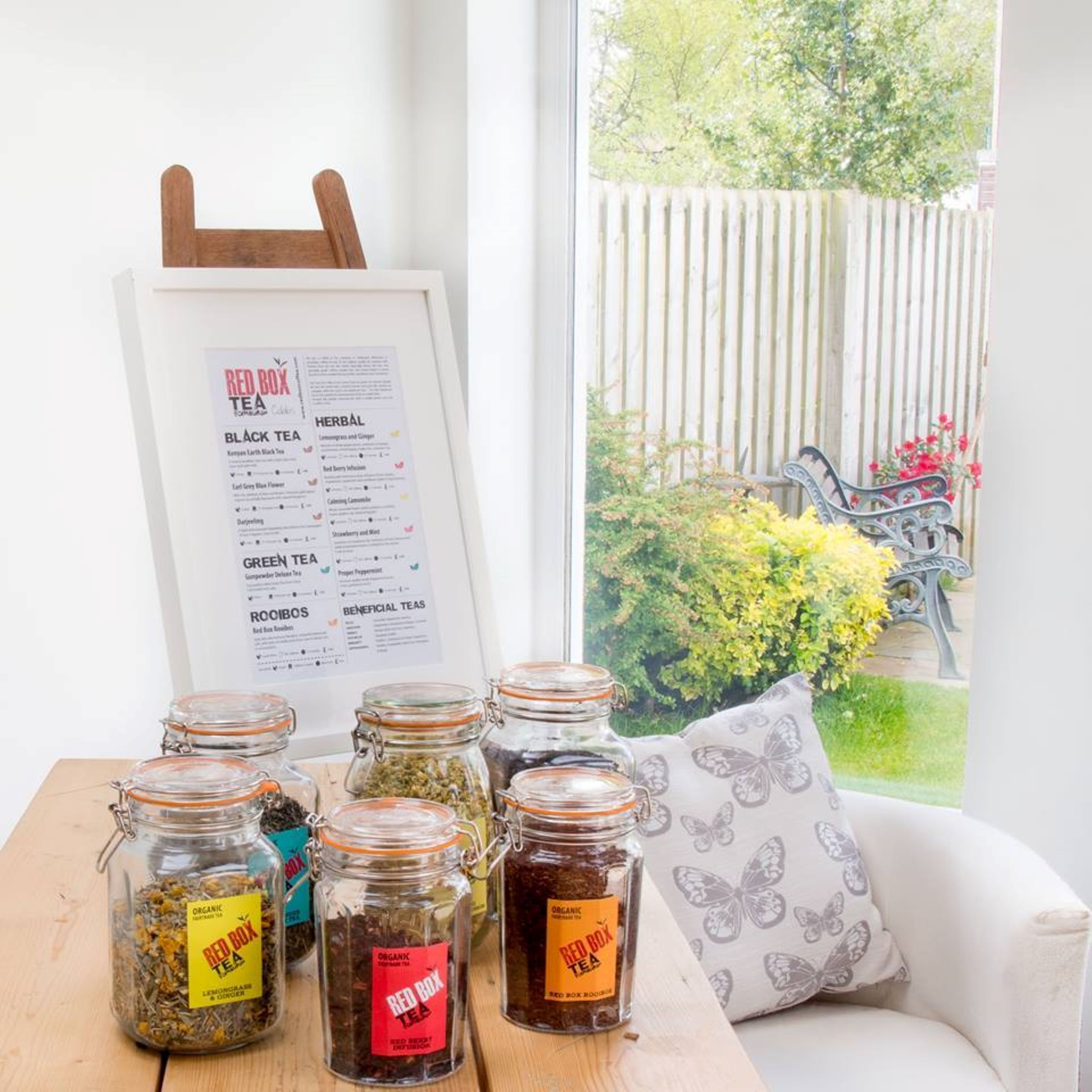 A menu board for Red Box Tea with various tea types listed, and several glass jars of tea on a table with a view of a garden outside.