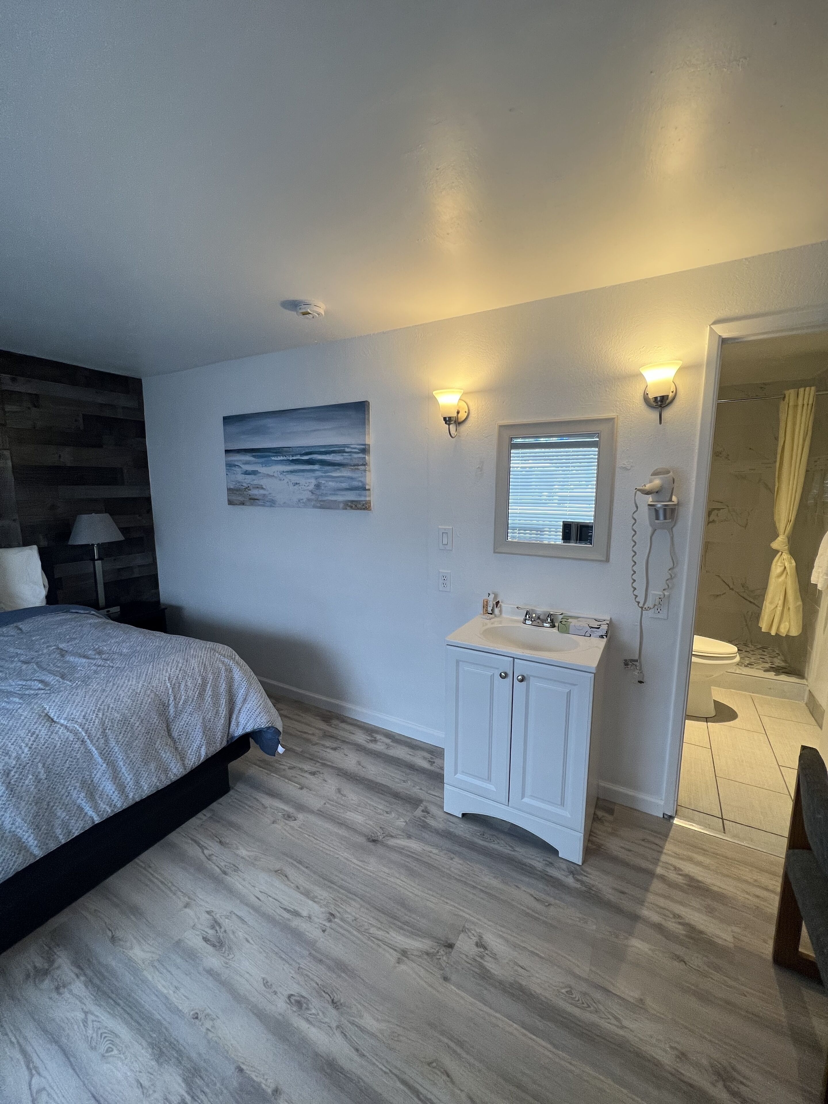 A bedroom with a bed, a nightstand, a wall-mounted lamp, a painting, a vanity with a mirror, and a shower area.