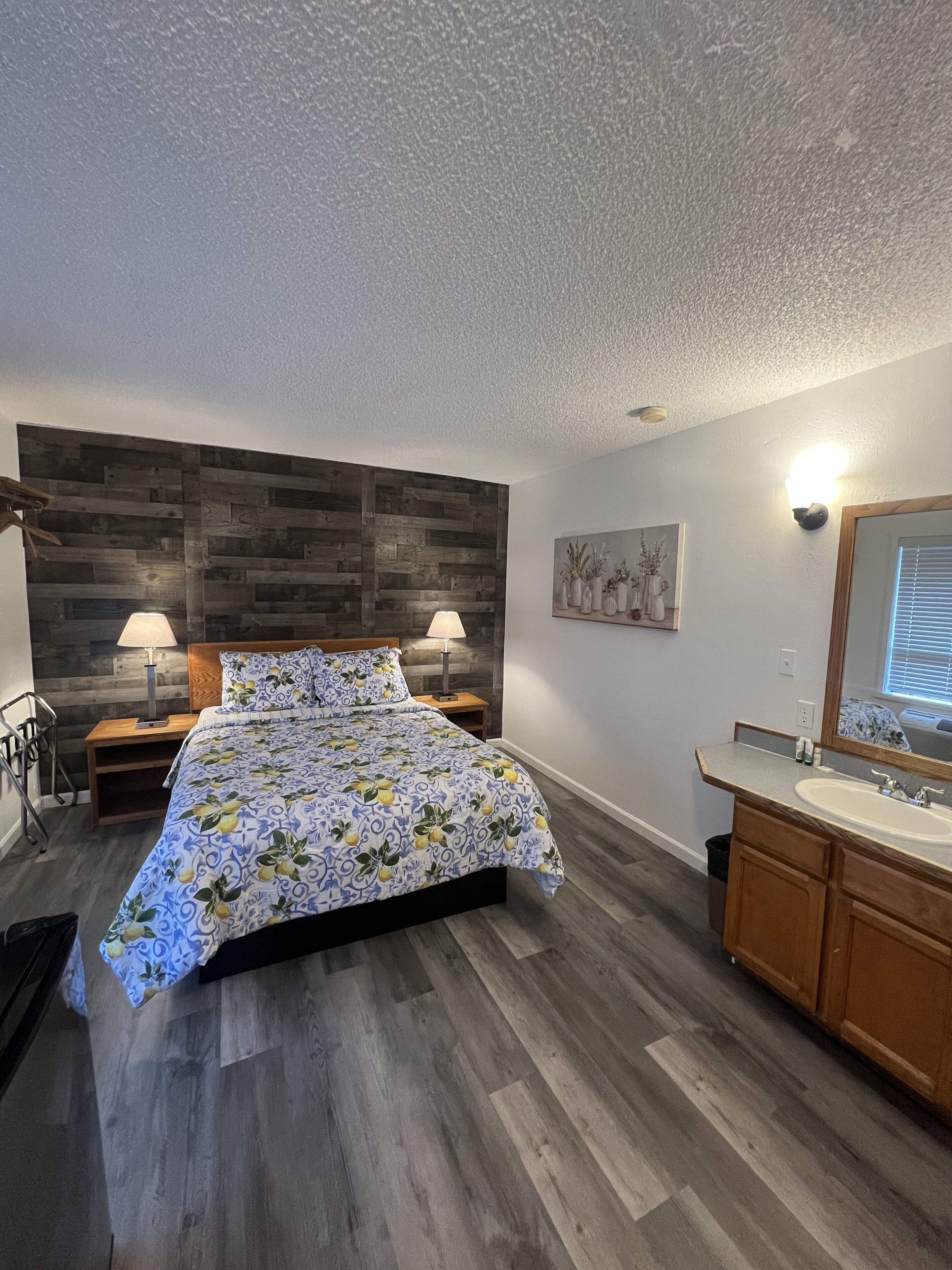 A bedroom with a wooden headboard, a bed with a floral bedspread, two bedside lamps, a nightstand, a wooden dresser with a mirror, and a small wall-mounted shelf with decorative items.
