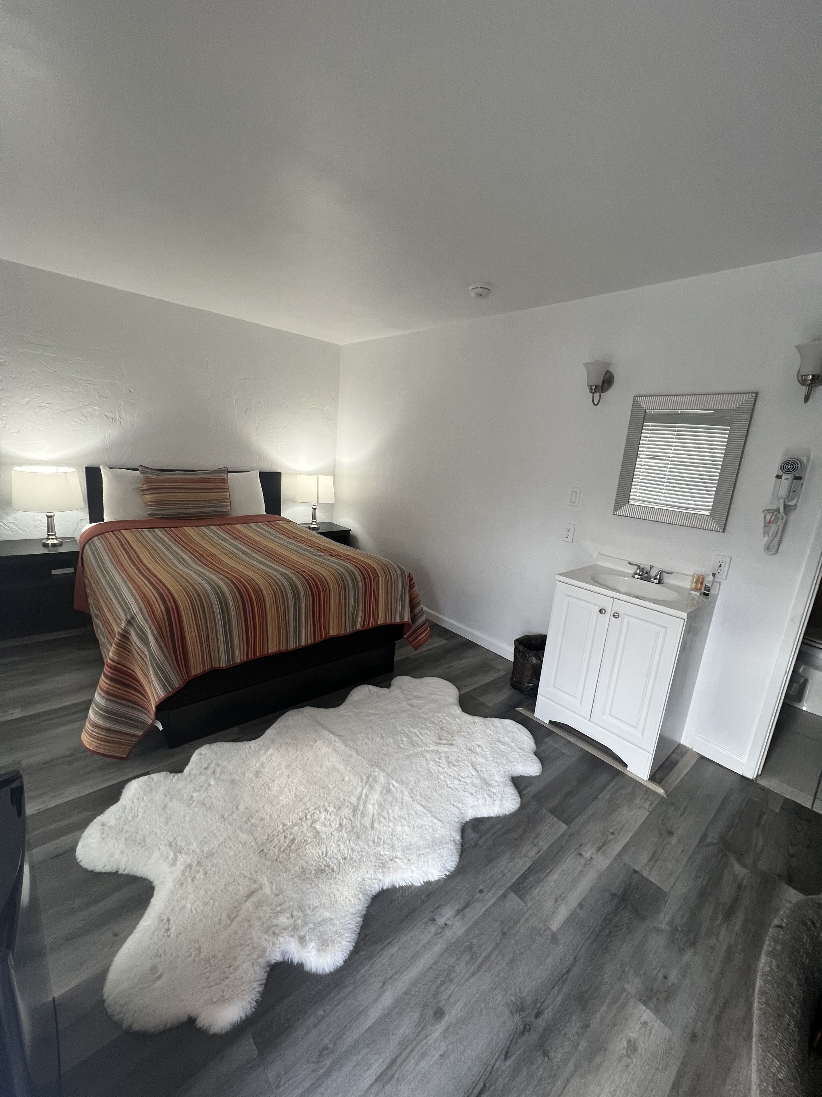 A bedroom with a bed, bedside lamps, a white rug, a vanity with a mirror, and a hairdryer.