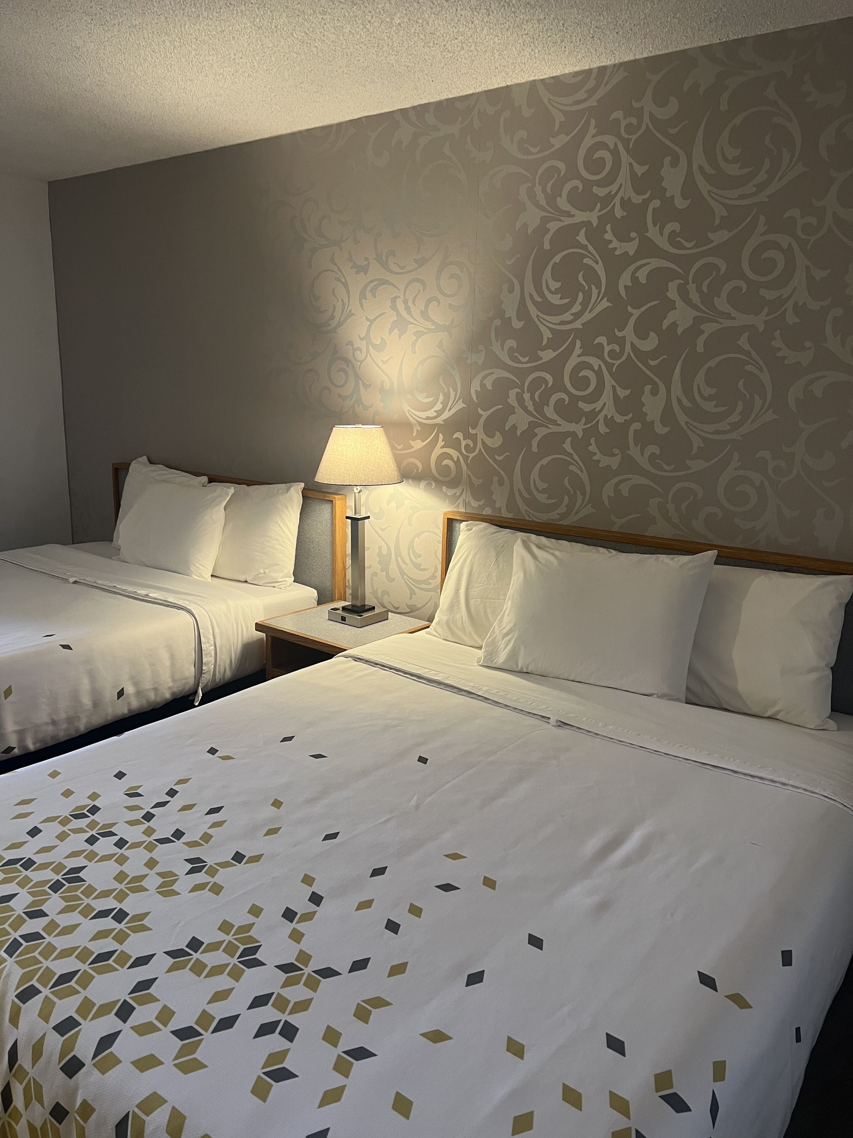 A hotel room with two beds, a nightstand with a lamp, and a patterned wallpaper.