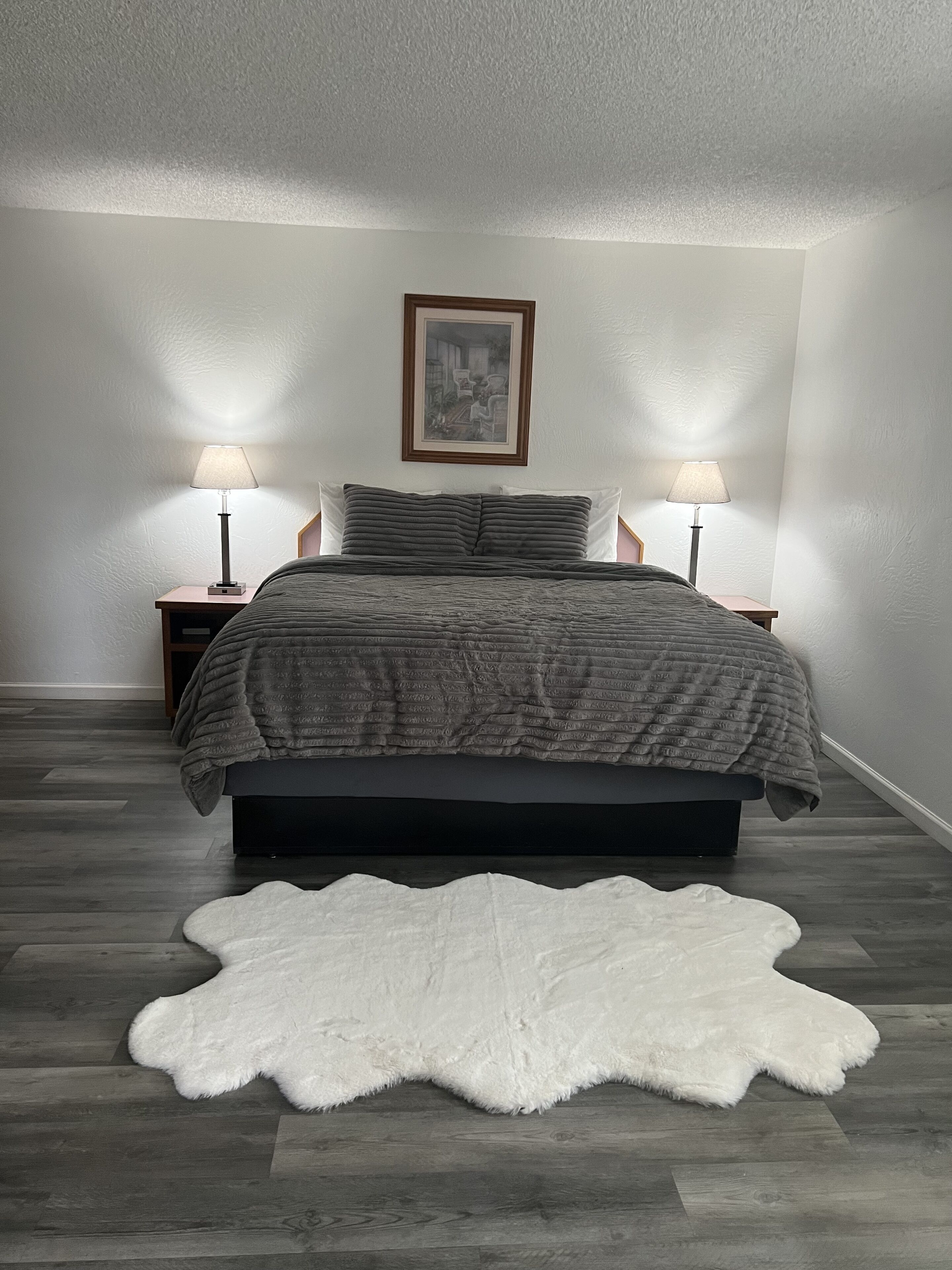 A bedroom with a bed, two bedside lamps, a framed picture on the wall, and a white furry rug on the floor.