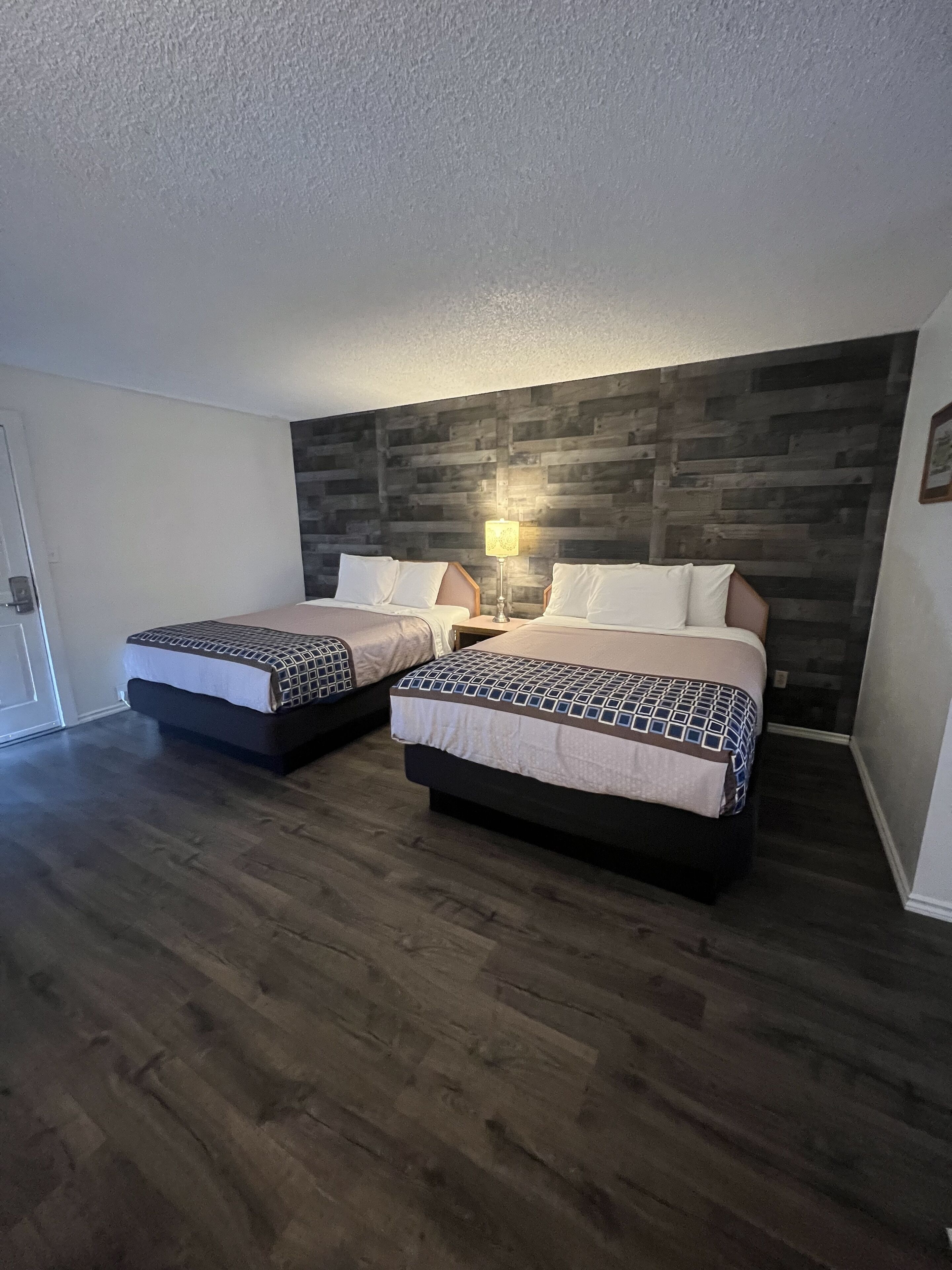 A hotel room with two beds, a wooden headboard, a nightstand with a lamp, and a door.