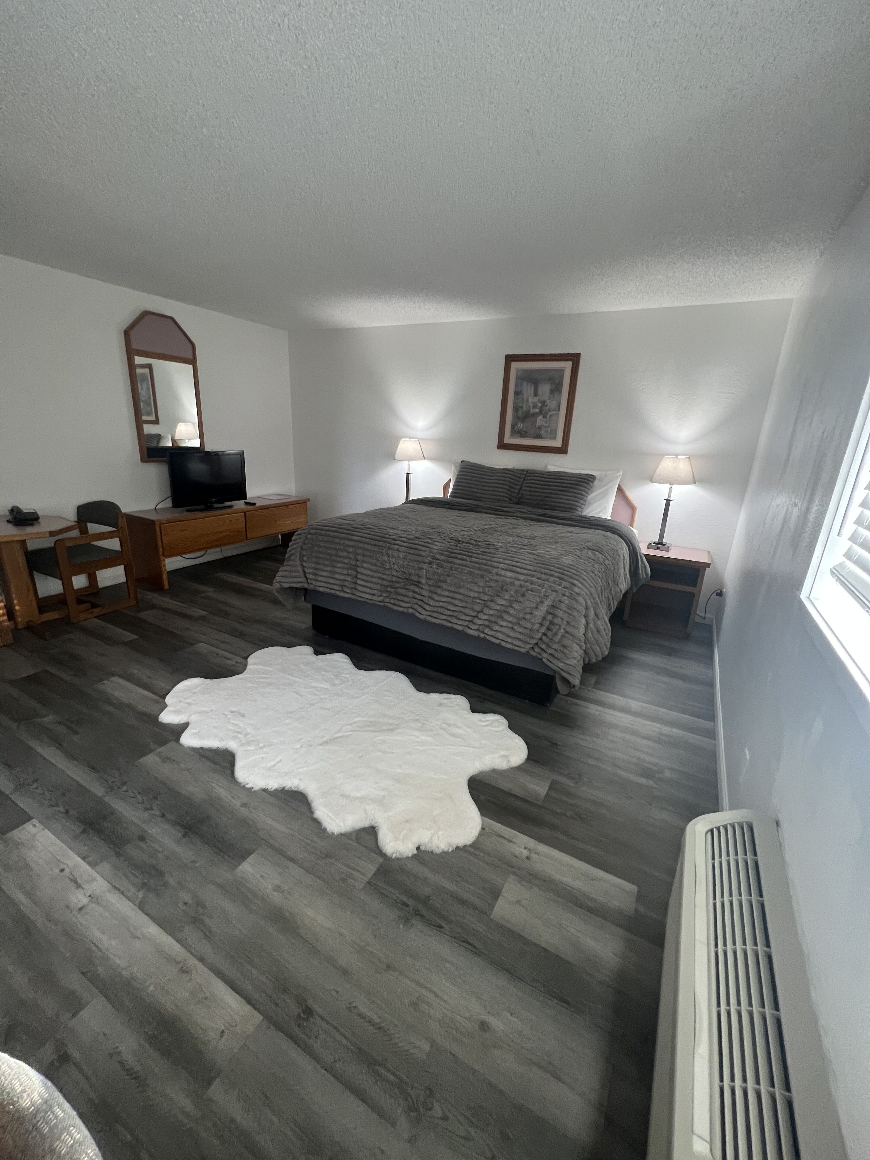 Een slaapkamer met een bed, een bureau, een stoel, een televisie, twee bedlampjes, een lijst met foto en een witte vloerkleed.