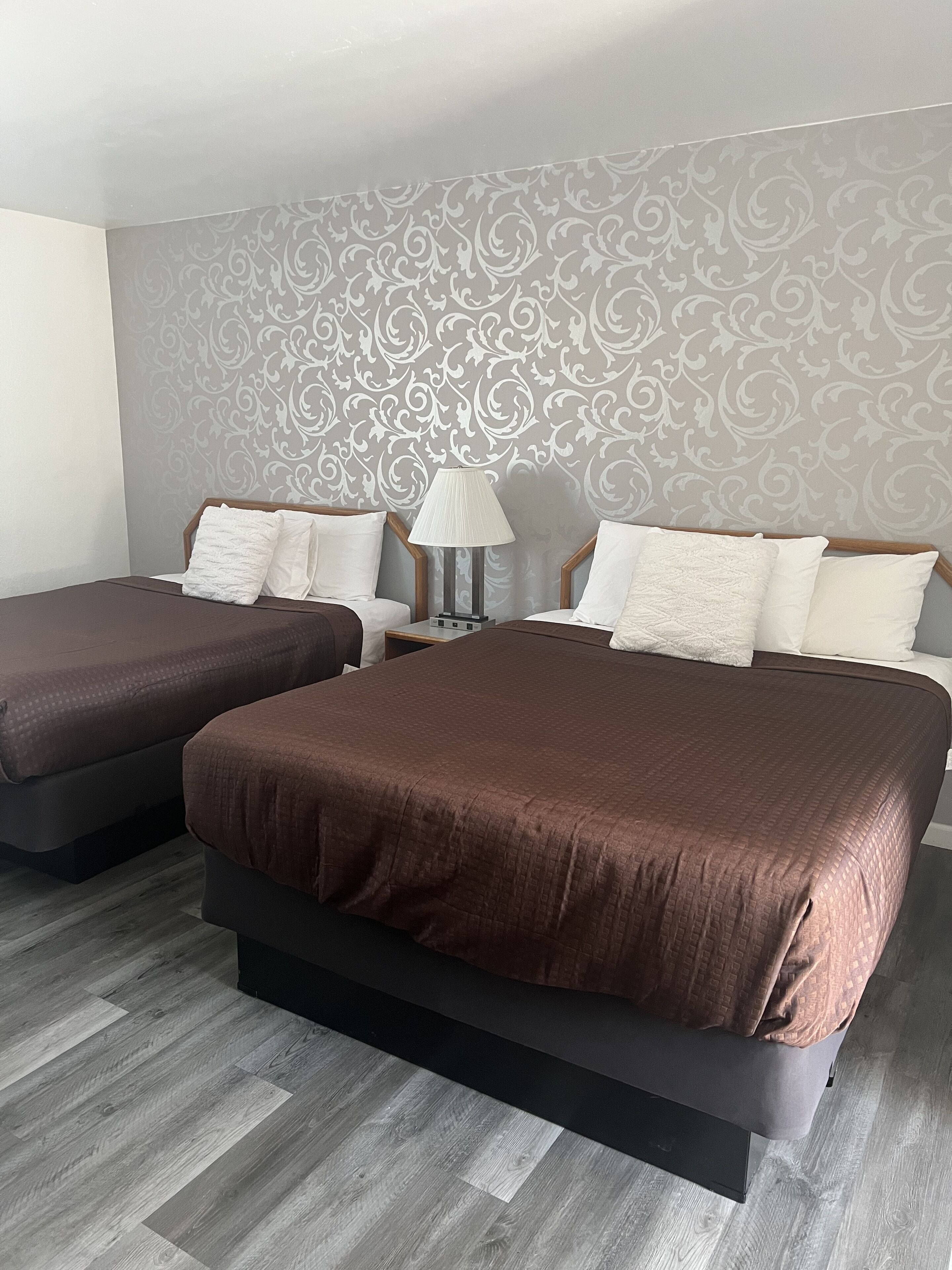 A hotel room with two beds, a nightstand with a lamp, and a patterned wallpaper.