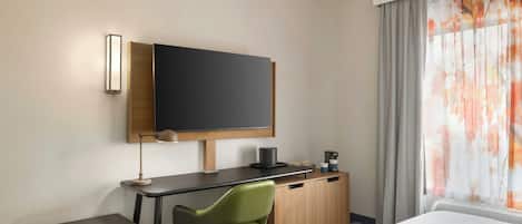 A hotel room with a bed, a desk with a chair, a flat-screen TV, a coffee maker, and a window with curtains.