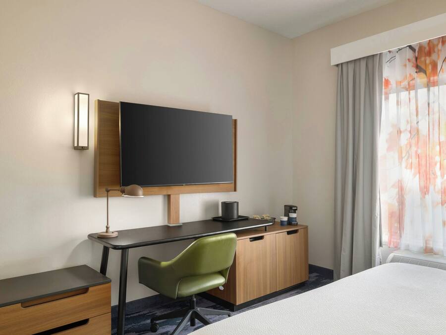 A hotel room with a bed, a desk with a chair, a flat-screen TV, a coffee maker, and a window with curtains.