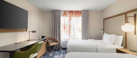 A hotel room with a desk, two beds, a chair, a TV, and a window with curtains.