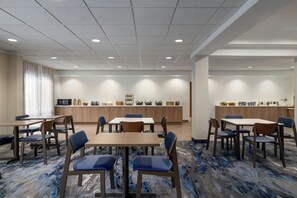 Une salle à manger d’hôtel avec plusieurs tables et chaises, un espace cuisine avec un buffet, et une grande fenêtre avec des rideaux.
