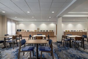 Un comedor de hotel con varias mesas y sillas, una zona de cocina con bufé, y un ventanal con cortinas.