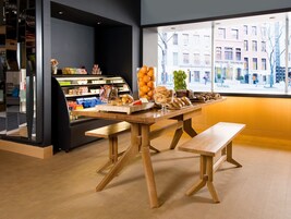 Un café con mesas y bancos de madera, una exposición de pasteles y vistas a la ciudad a través de amplias ventanas.