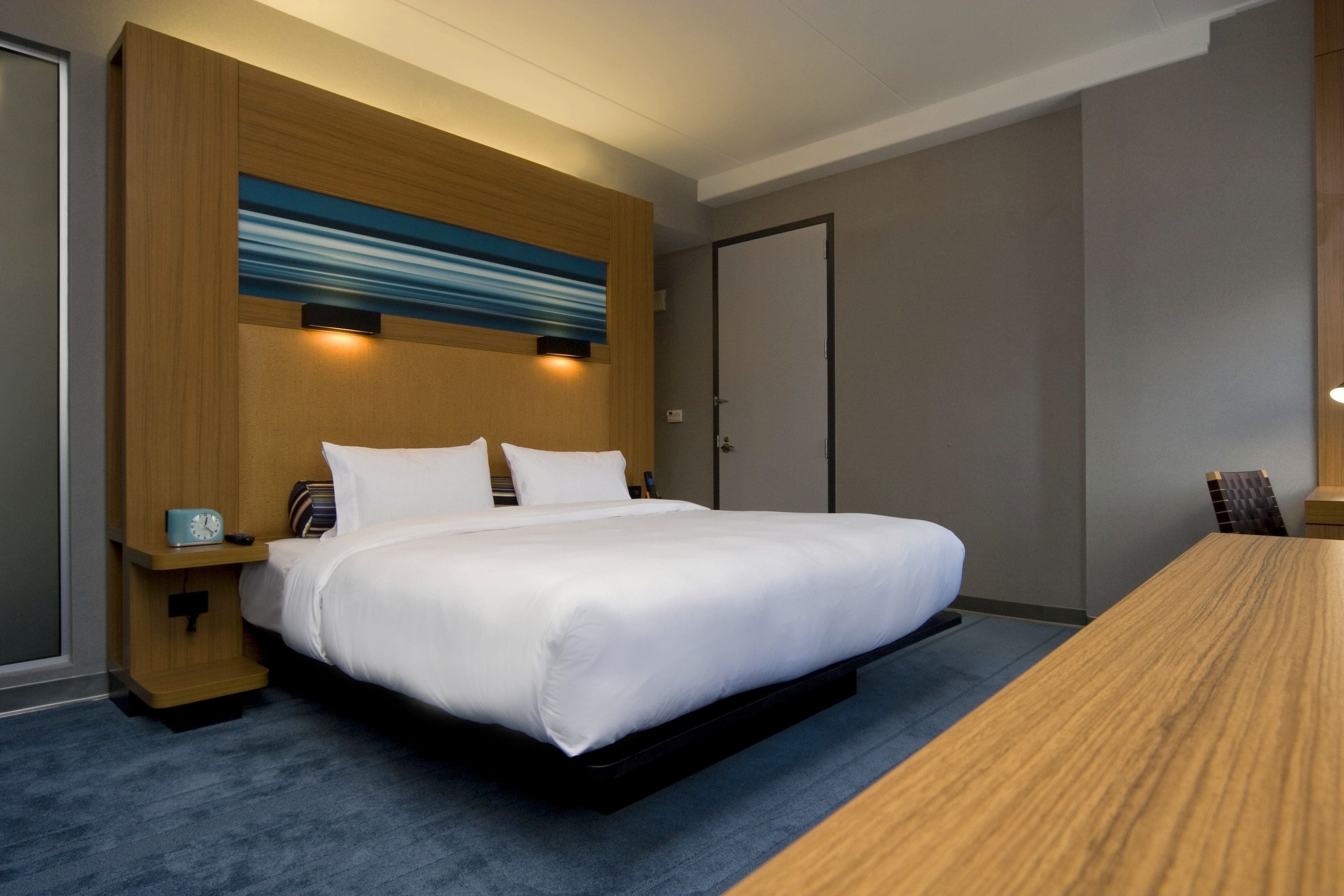 A hotel room with a large bed, a wooden headboard, a blue abstract painting, a bedside table with a clock, and a wooden desk.