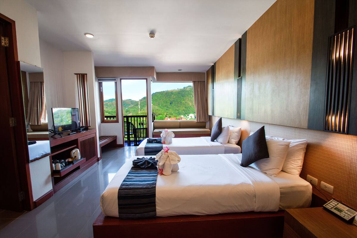 A hotel room with a large bed, a television, and a balcony with a view of greenery.
