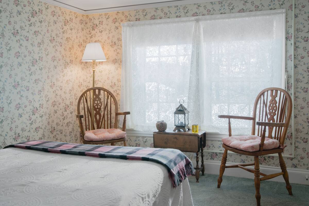 A bedroom with floral wallpaper, a bed with a quilt, two wooden chairs, a nightstand with a lamp, and a window with a lace curtain.