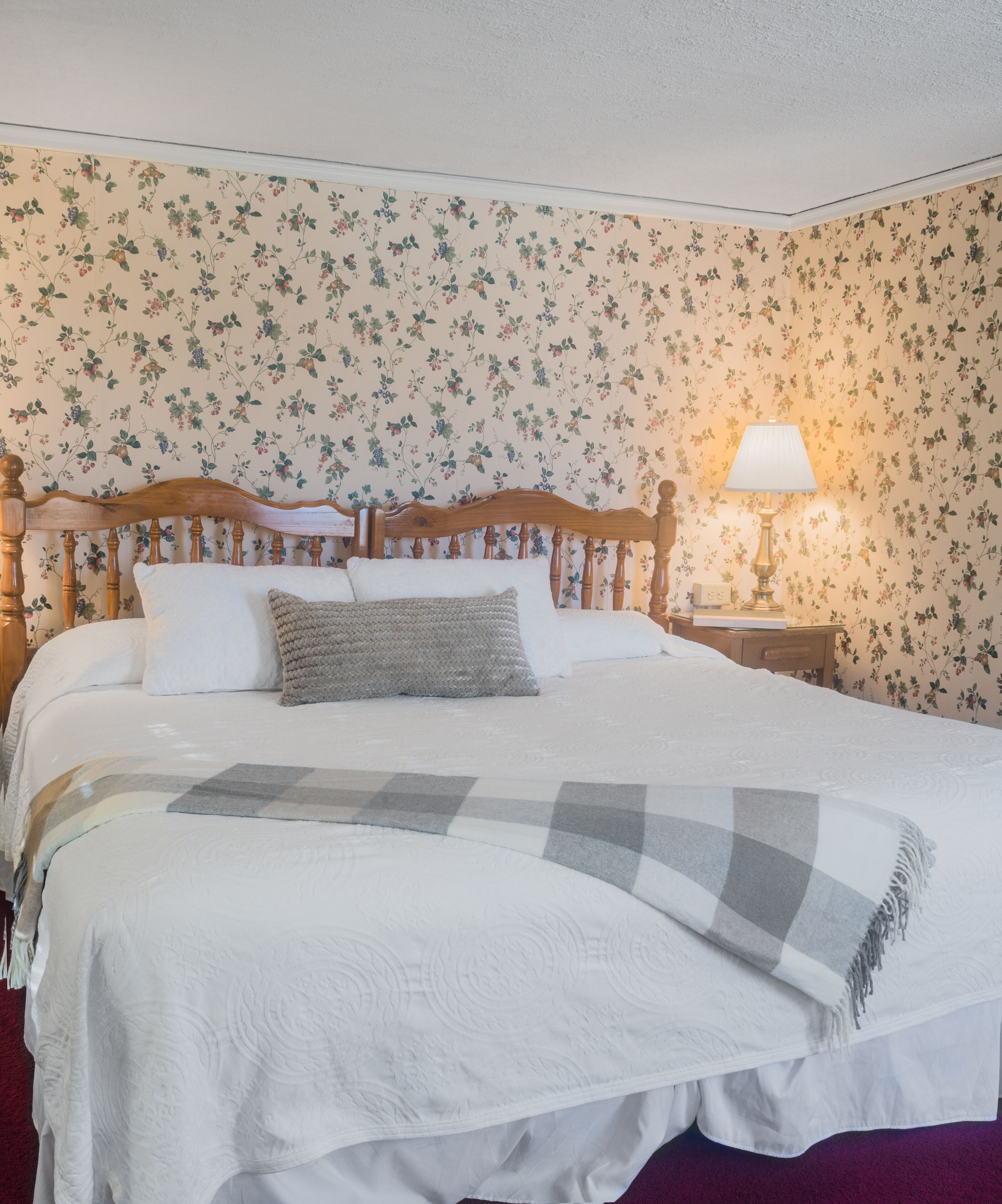 Cama bem arrumada com manta xadrez, cabeceira de madeira e papel de parede floral.