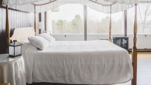 A four-poster canopy bed with a white bedspread, a bedside table with a lamp, and a view of trees through large windows. - Dexter's Inn (Sunapee)