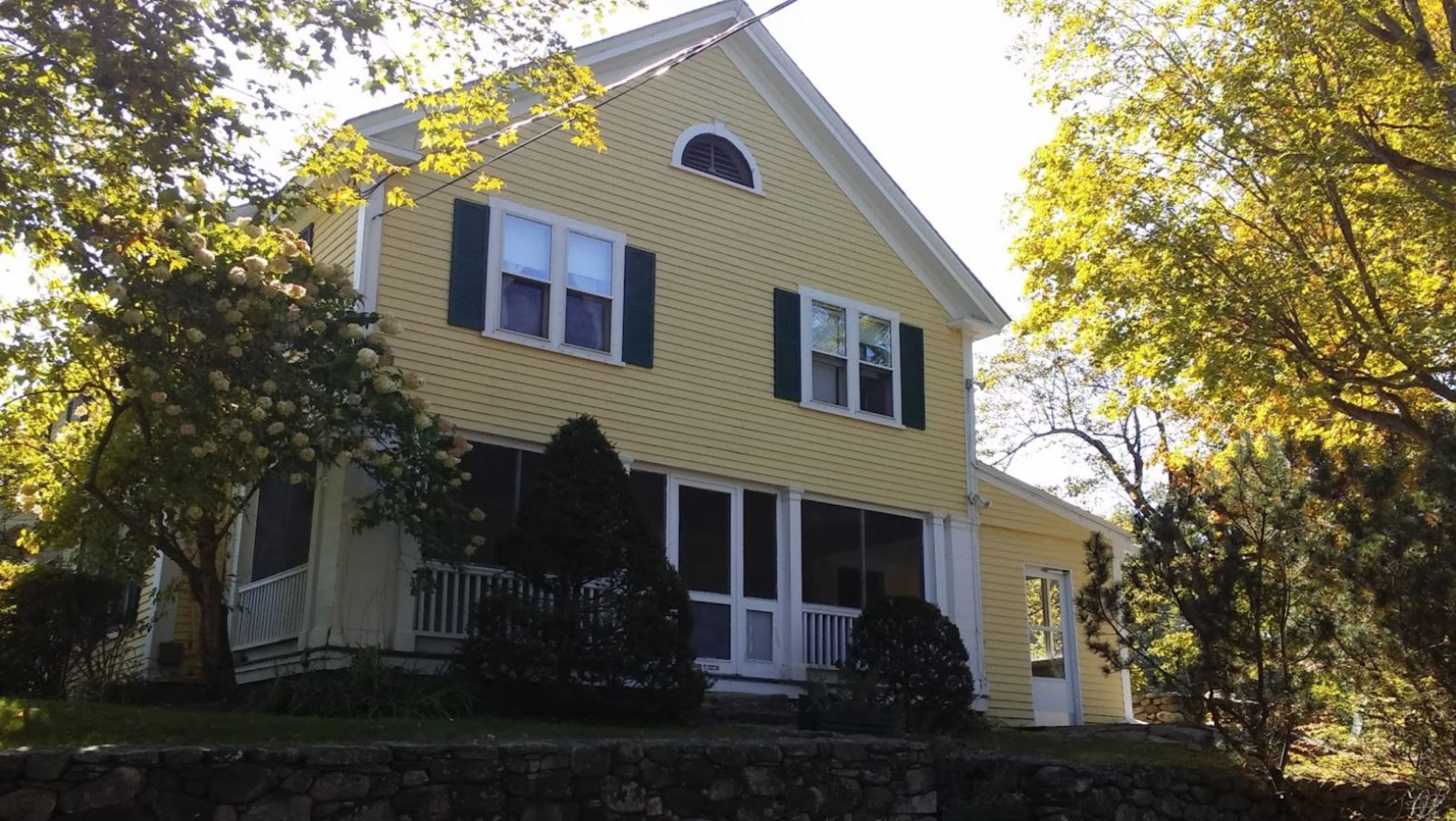 A two-story house with a front porch, a stone wall in front, and trees with yellow leaves around.