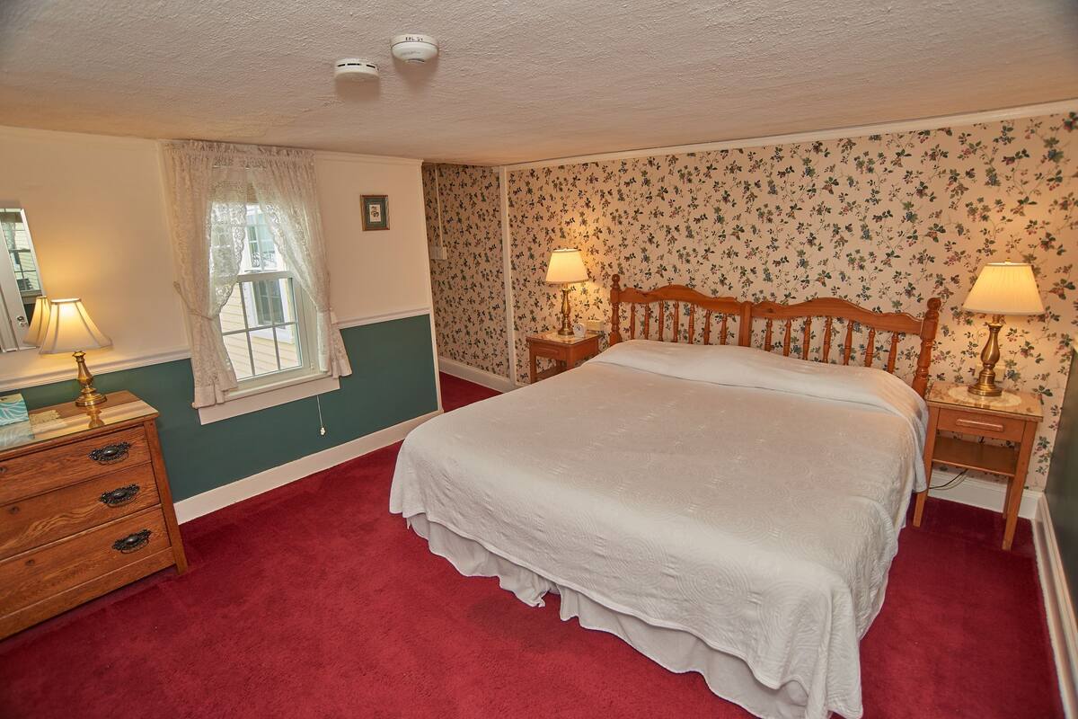 A bedroom with a large bed, two bedside tables with lamps, a wooden dresser, floral wallpaper, and a window with a sheer curtain.