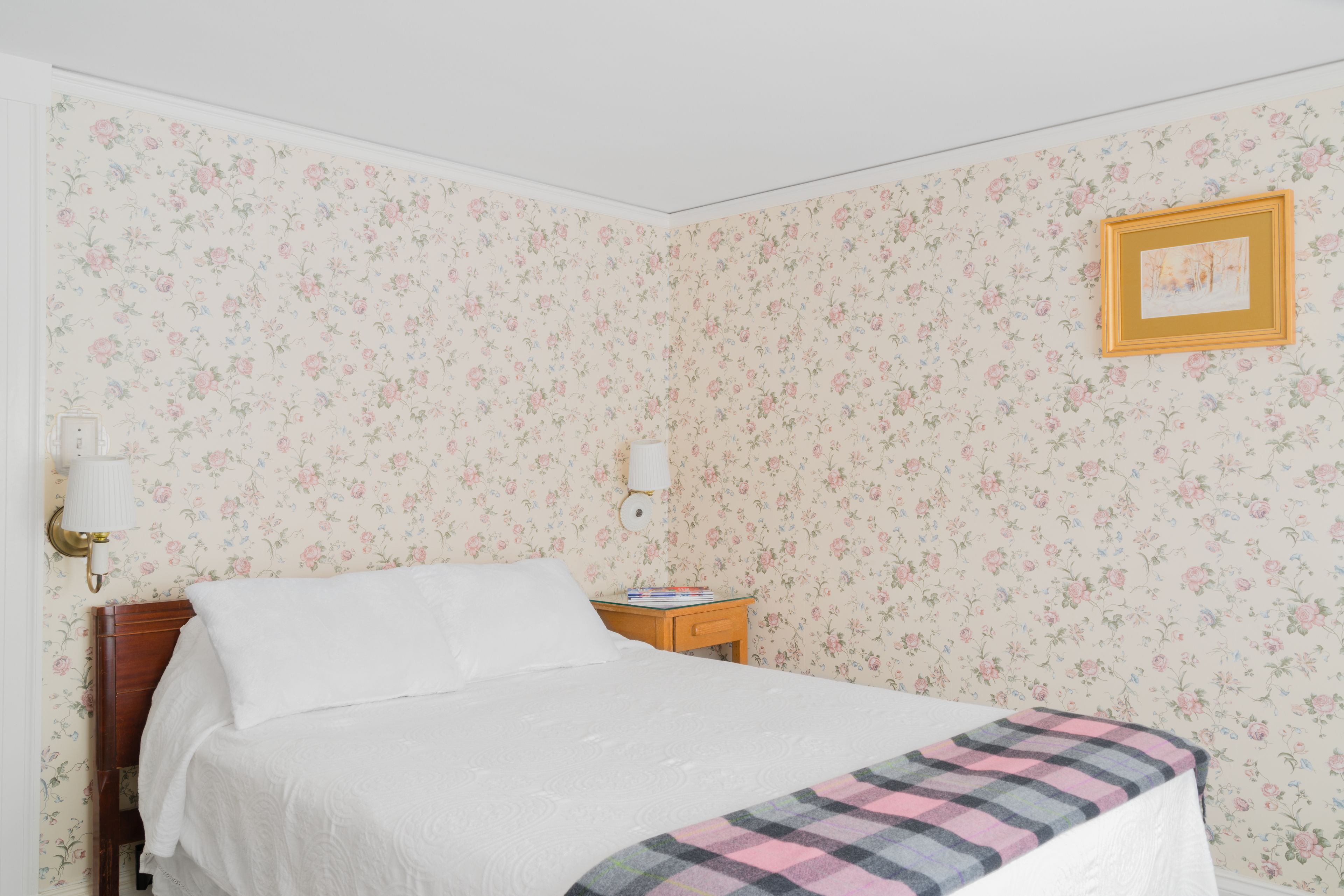 Ein ordentlich bezogenes Bett mit kariertem Überwurf, ein Nachttisch mit Büchern, eine Wandlampe und ein gerahmtes Bild an der Wand.
