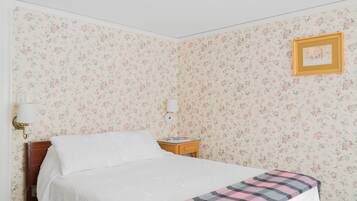 Ein ordentlich bezogenes Bett mit kariertem Überwurf, ein Nachttisch mit Büchern, eine Wandlampe und ein gerahmtes Bild an der Wand.