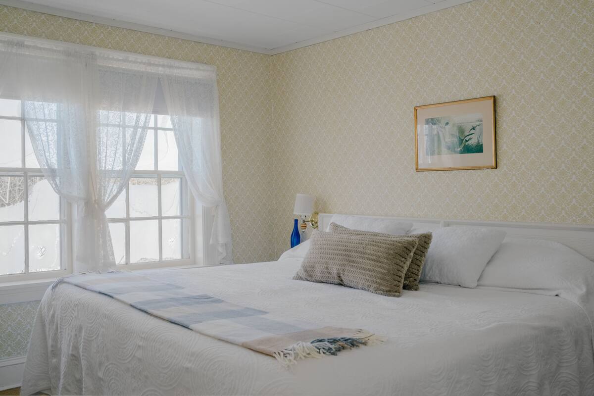 A neatly made bed with a quilt, a framed picture on the wall, and a window with sheer curtains.
