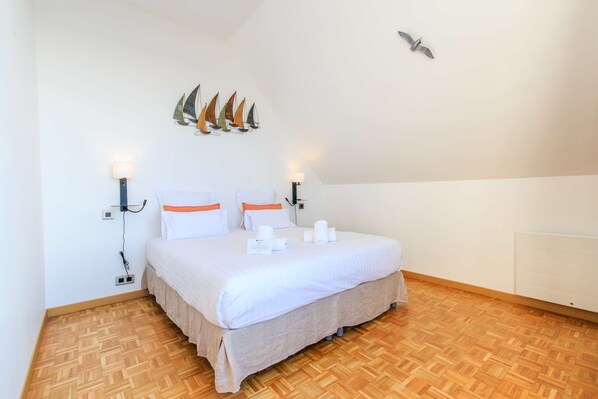 Una cama bien hecha con ropa blanca y detalles naranjas, suelo de parqué de madera y lámparas fijadas a la pared.