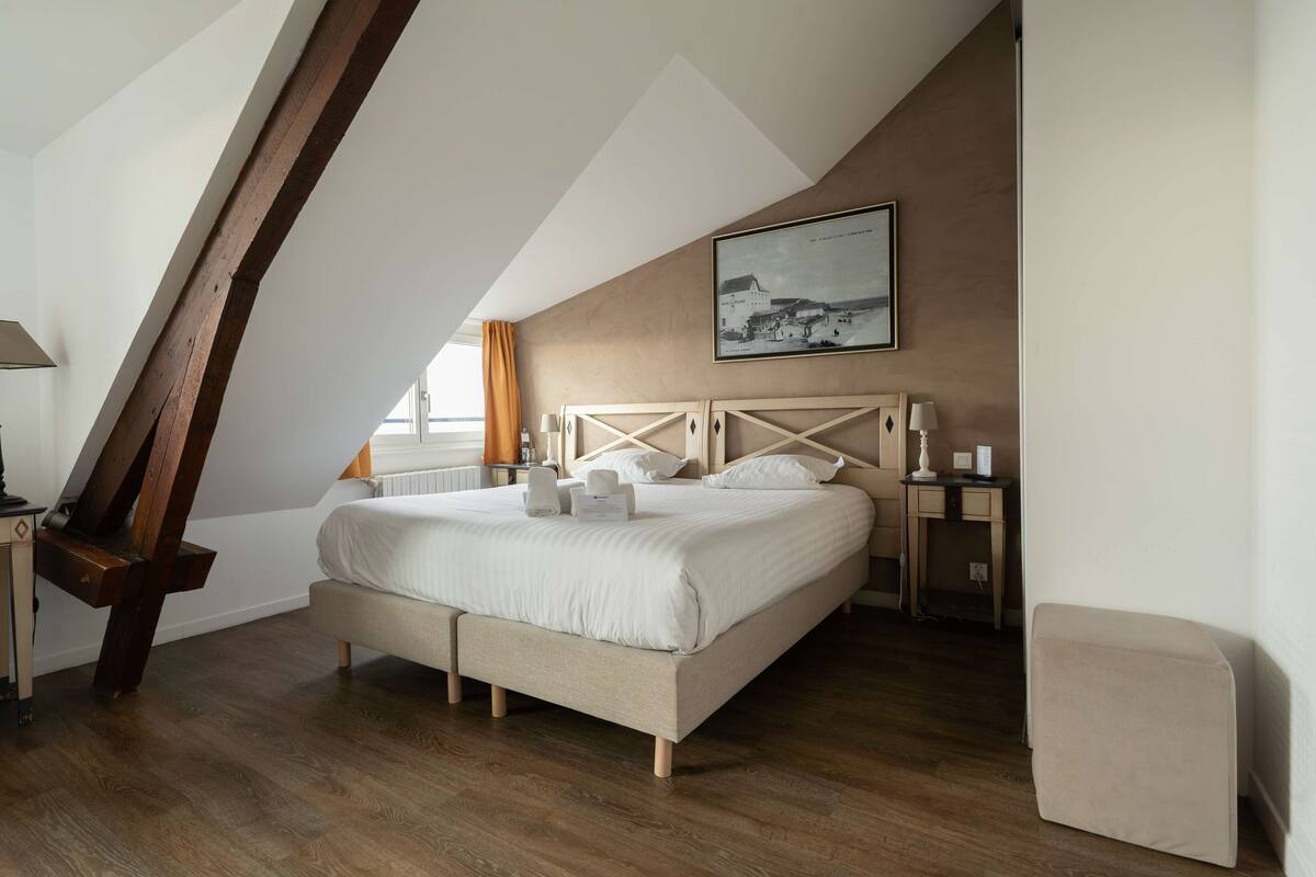 A bedroom with a sloped ceiling, a bed with white bedding, a wooden headboard, a bedside table with a lamp, and a framed picture on the wall.