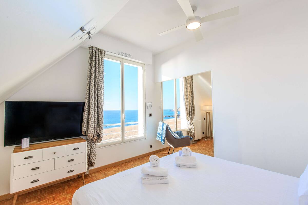 A bedroom with a TV, a bed with towels, a chair, and a view of the beach.