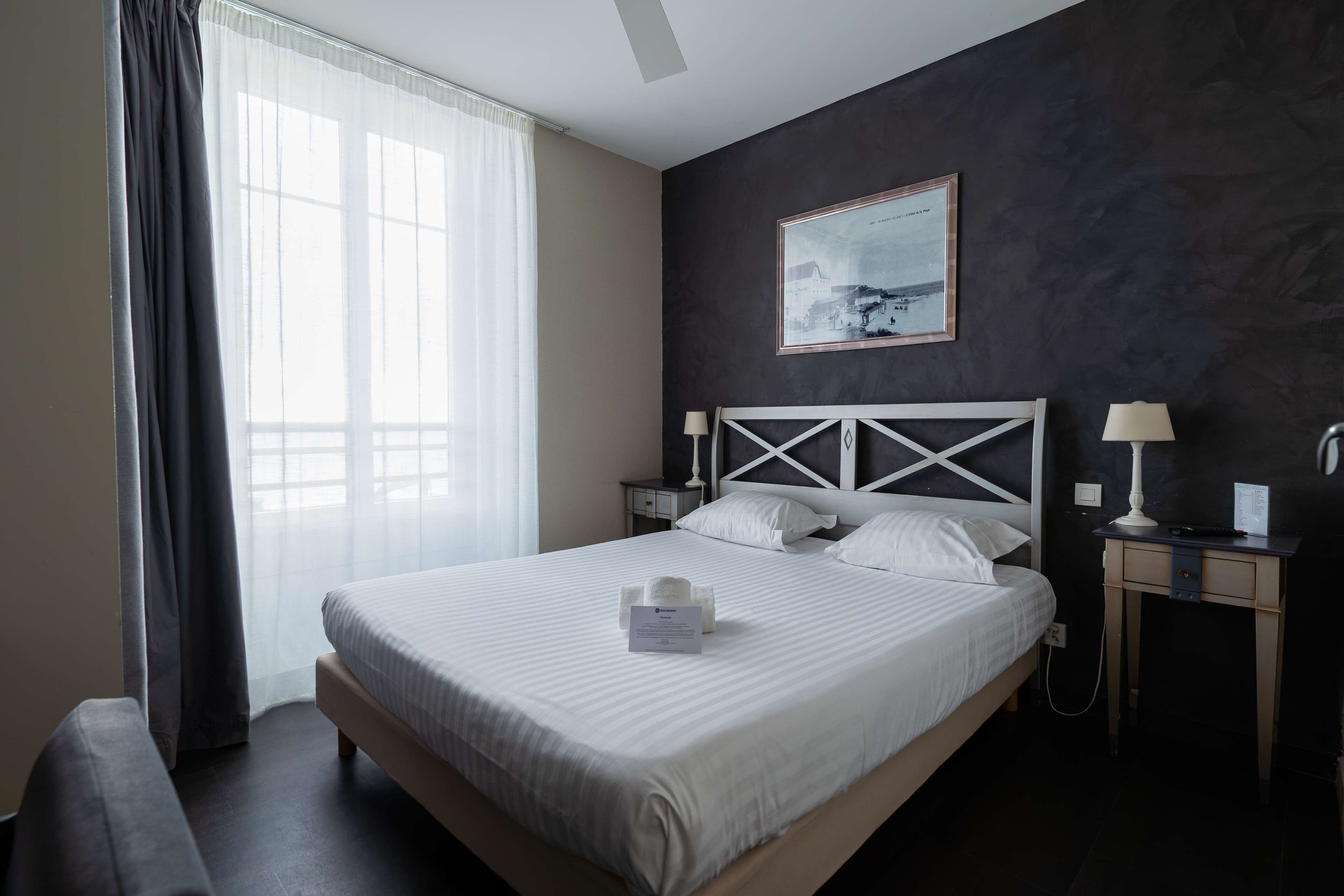 Une chambre d’hôtel avec un grand lit, une table de chevet avec une lampe, une tête de lit au motif géométrique, un tableau encadré au mur et une fenêtre avec des rideaux.