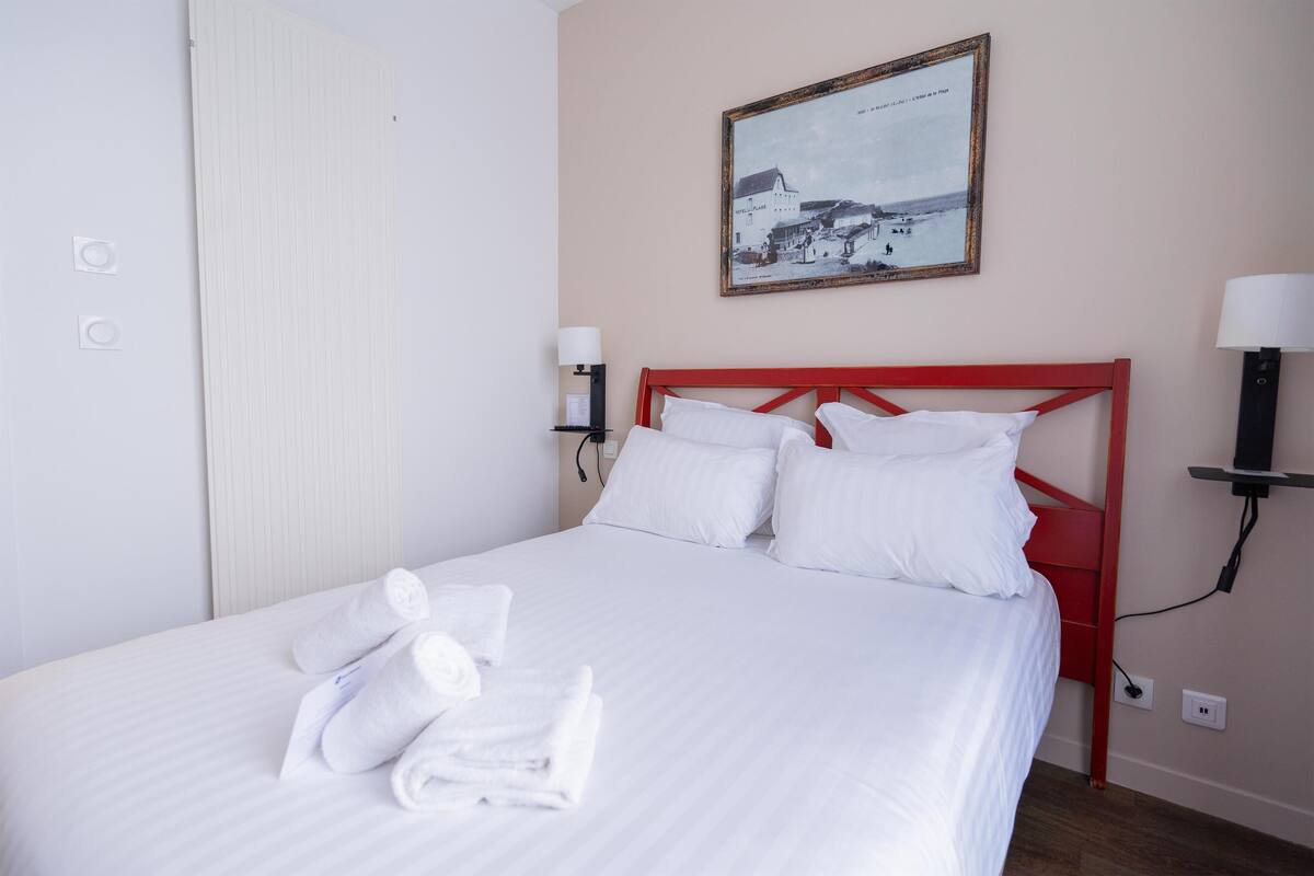 A neatly made bed with white linens and towels, a red headboard, and a framed picture on the wall.