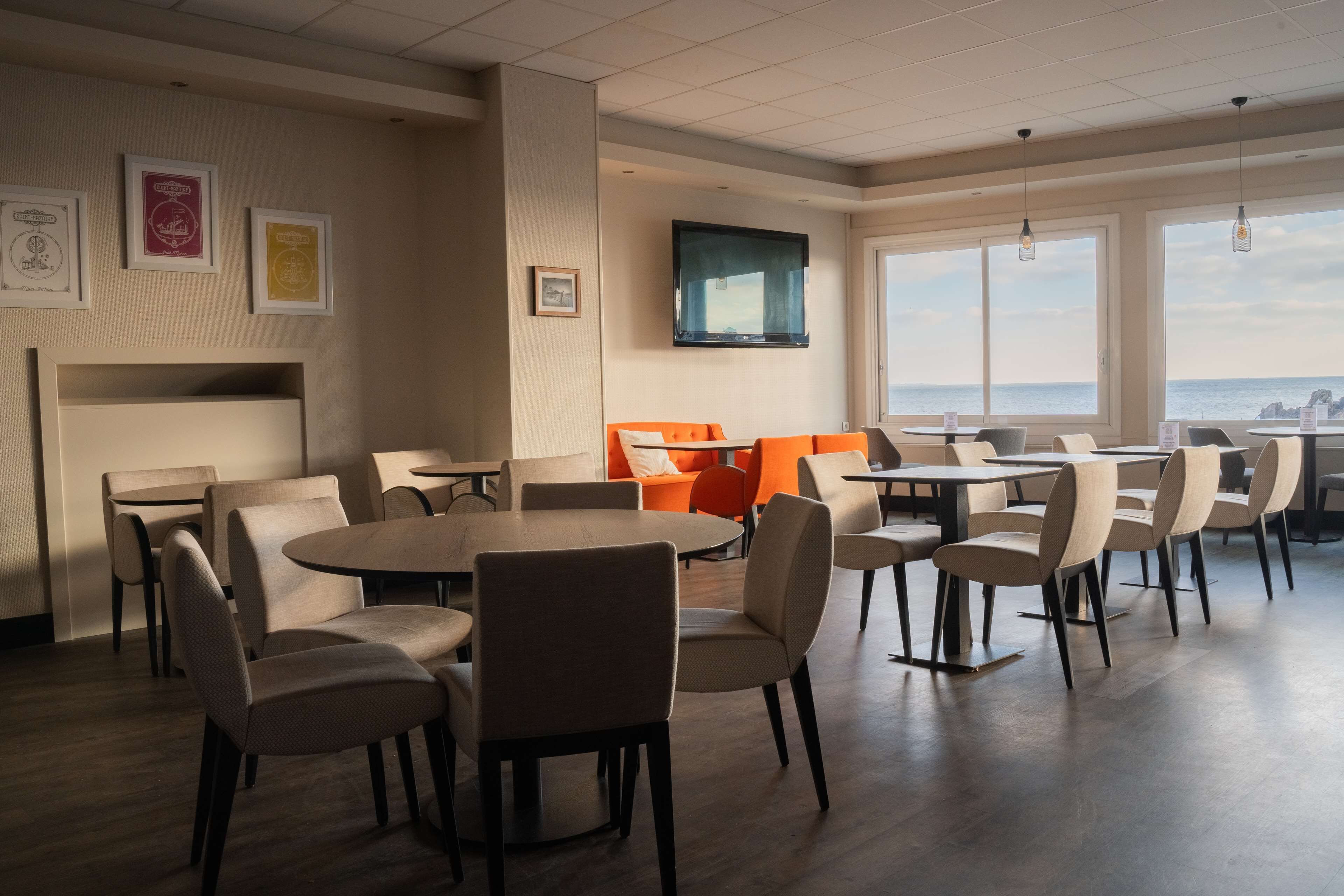 A dining area with round tables and chairs, a TV mounted on the wall, and a view of the sea through large windows.