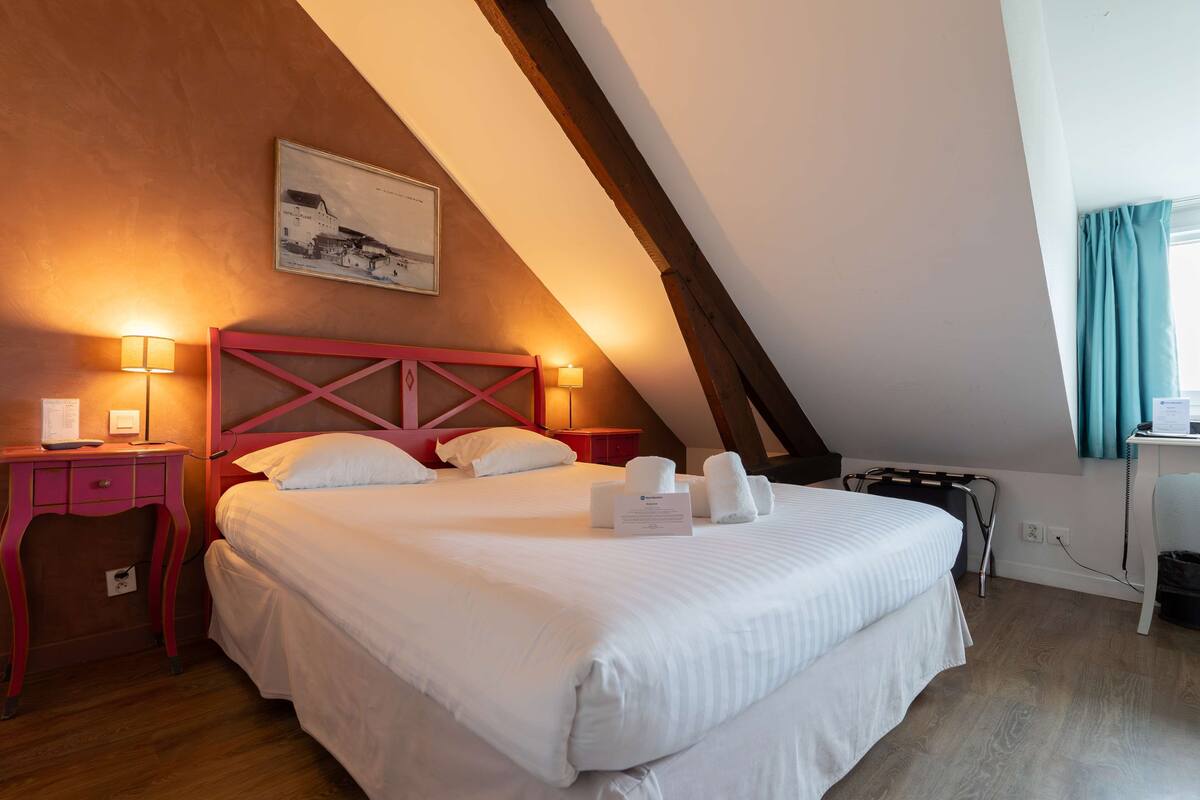 A neatly made bed in a room with a sloped ceiling, a wooden desk, and a wall-mounted lamp.