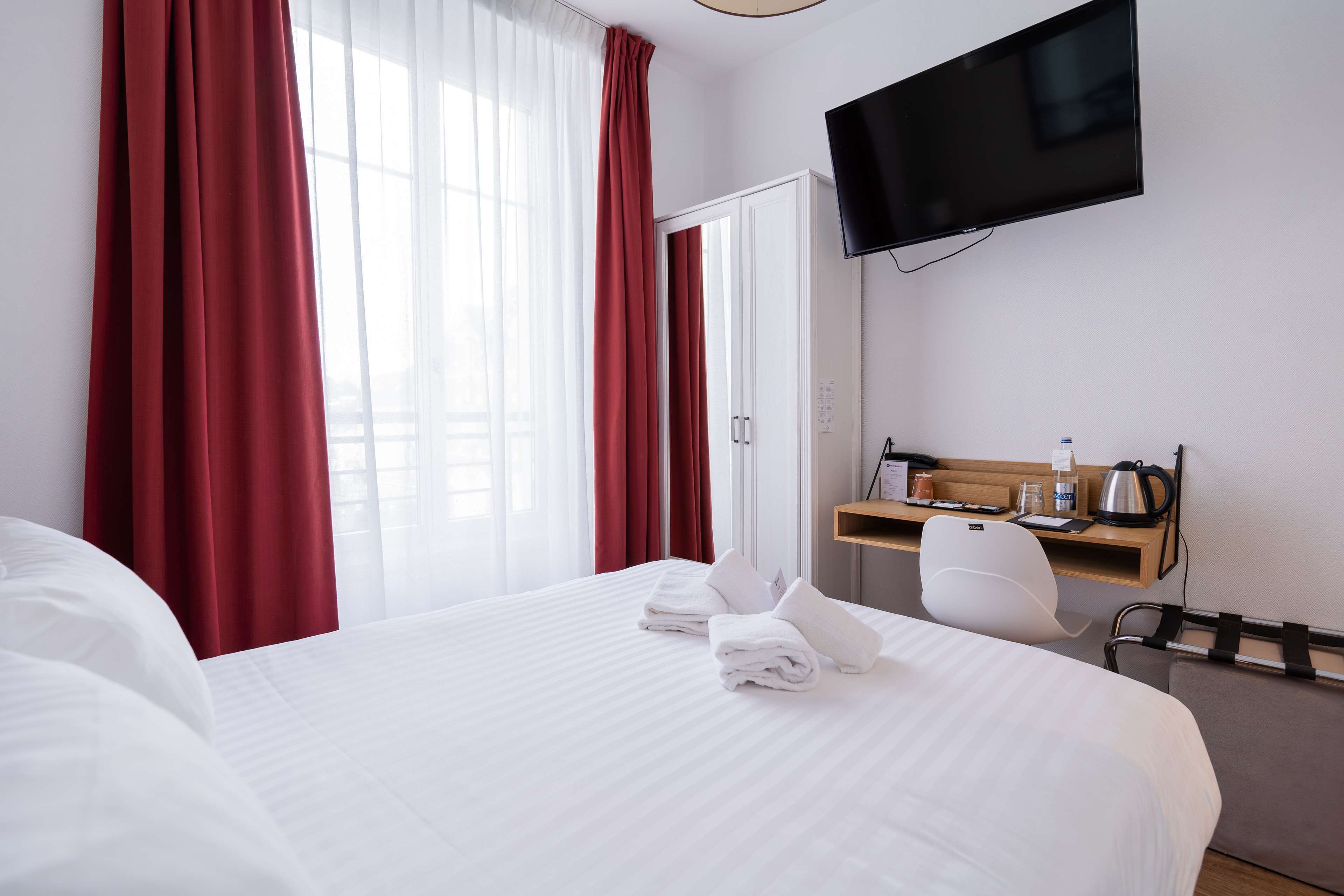 A hotel room with a large bed, a desk with a chair, a flat-screen TV, and a window with red curtains.