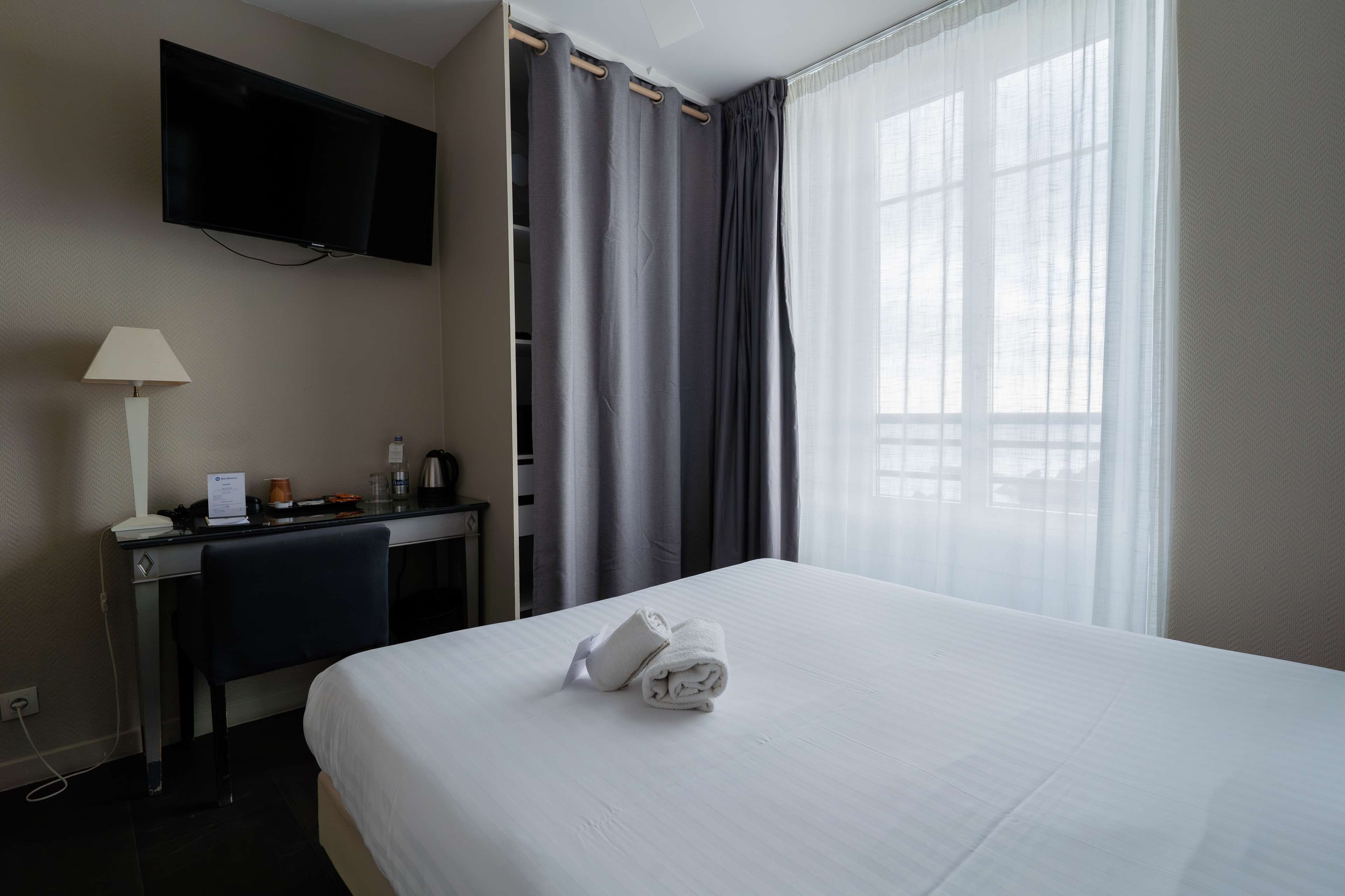 Une chambre d’hôtel avec un lit, un bureau doté d’une lampe, un téléviseur fixé au mur et une fenêtre avec des rideaux.
