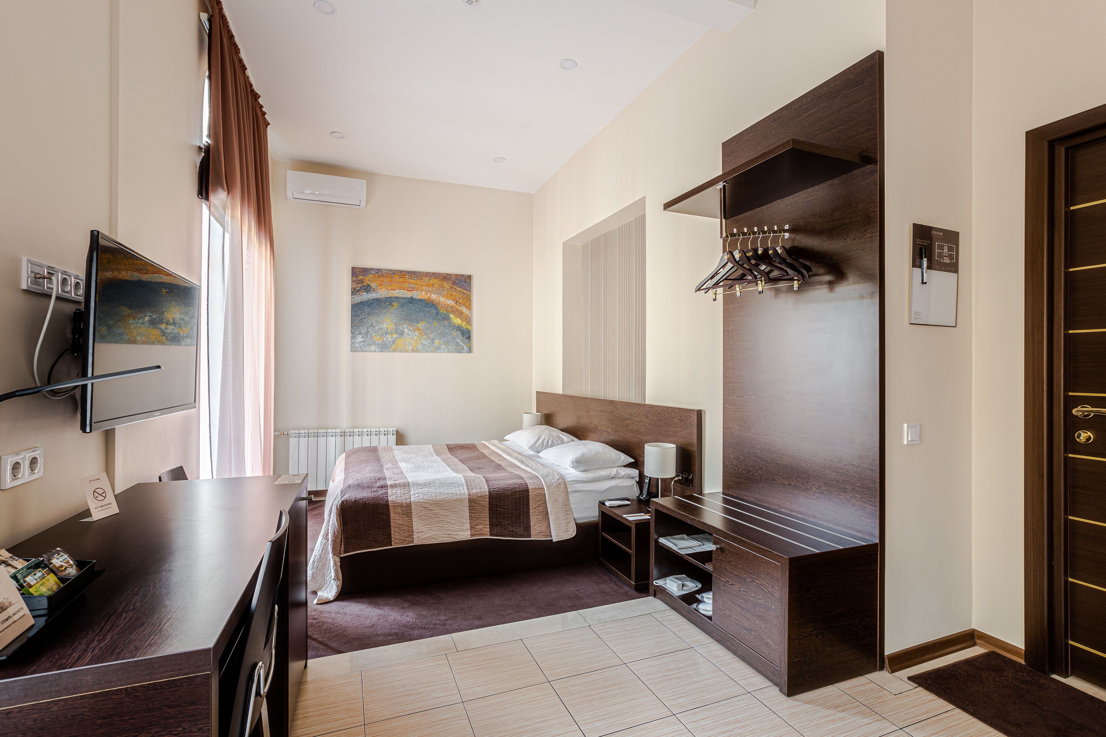 Habitación de hotel con cama, escritorio, televisión, un cuadro y perchero.