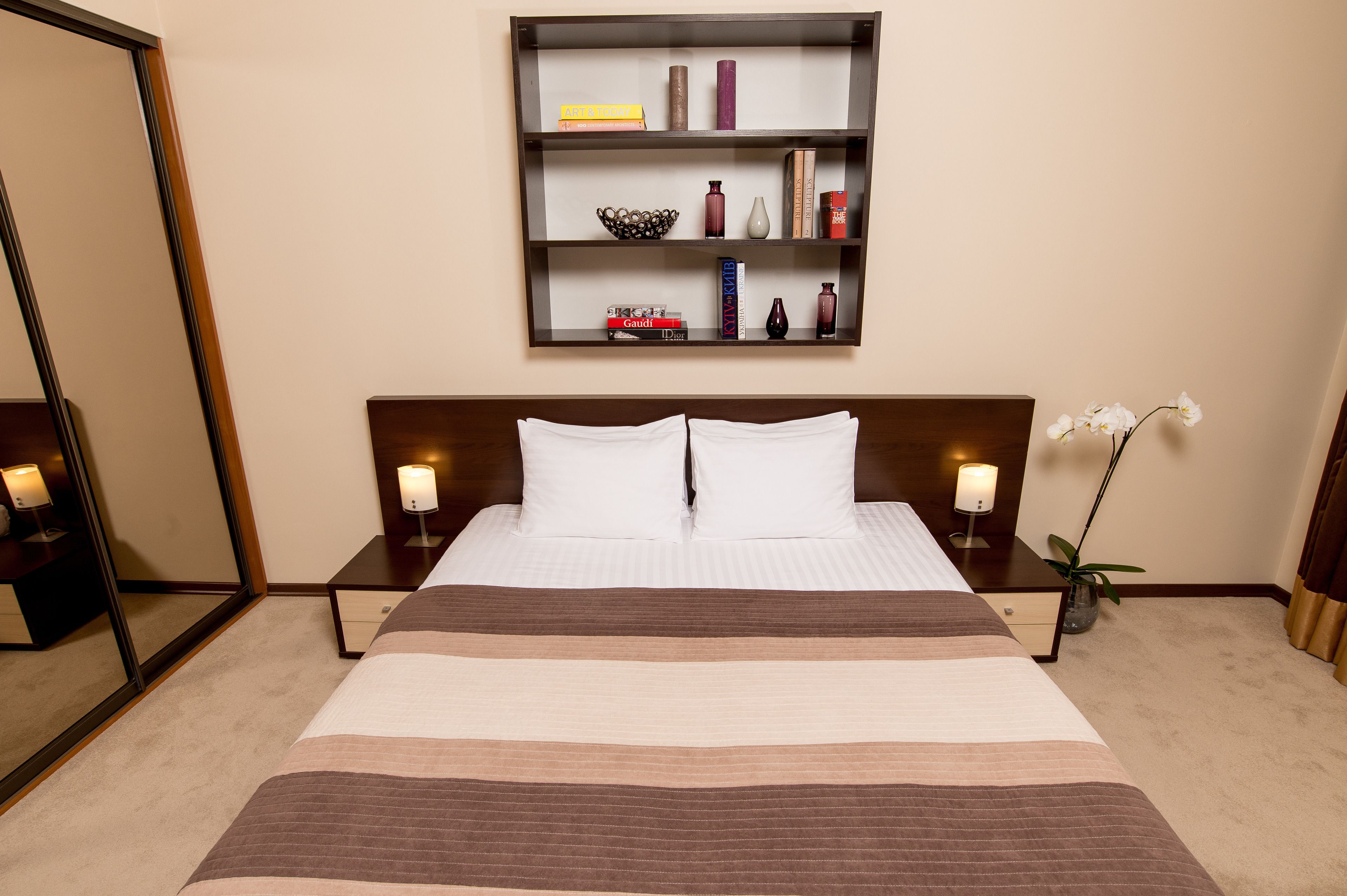 A hotel room with a large bed, two bedside tables with lamps, a wall-mounted shelf with books and decorative items, and a mirror.