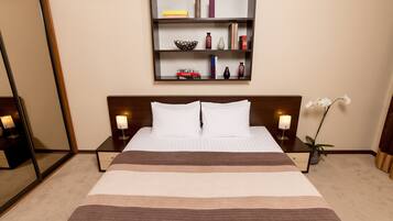 A hotel room with a large bed, two bedside tables with lamps, a wall-mounted shelf with books and decorative items, and a mirror.
