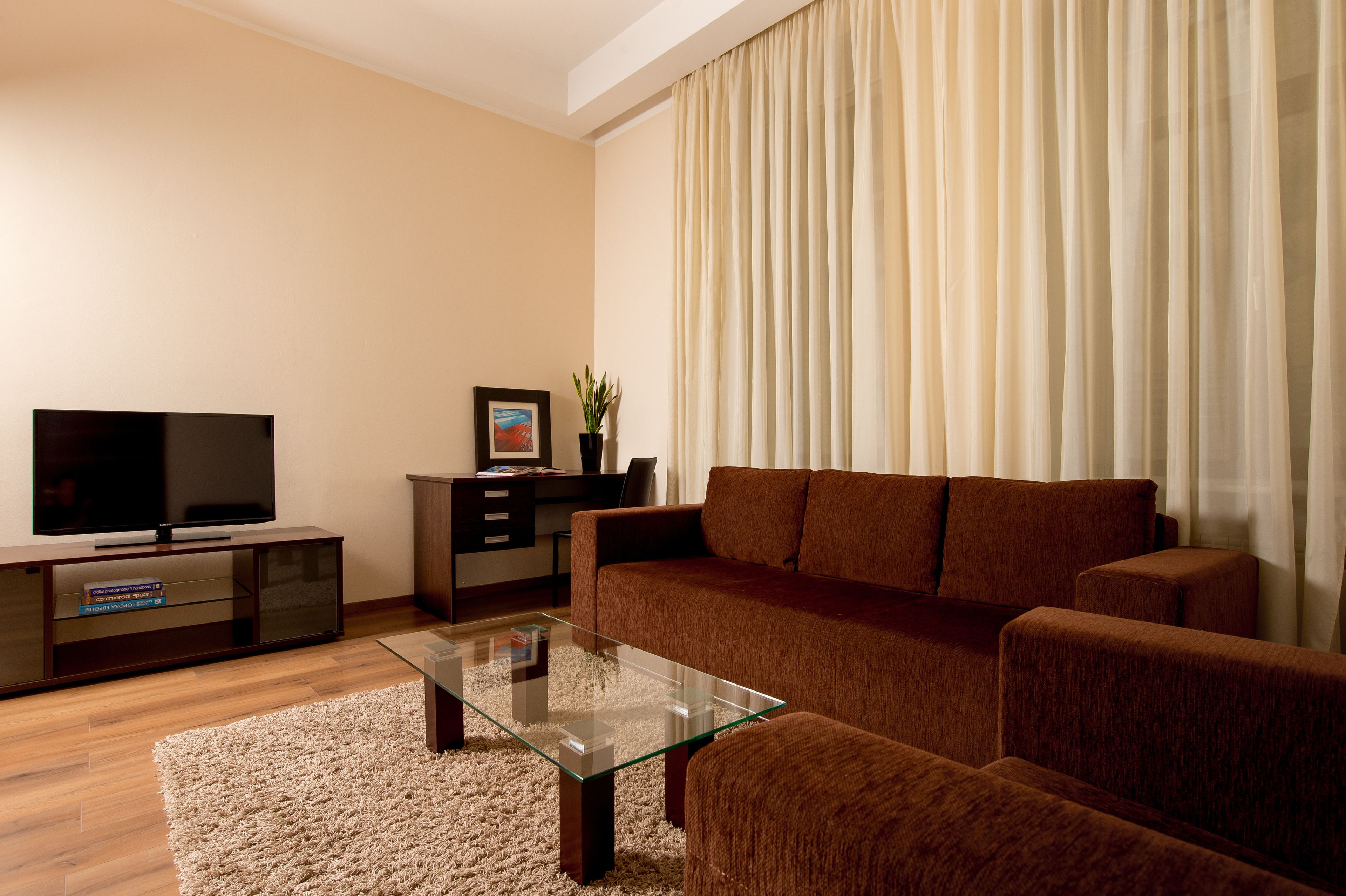 A modern living room with a flat-screen TV, a glass coffee table, a brown sofa, and a large window with curtains.