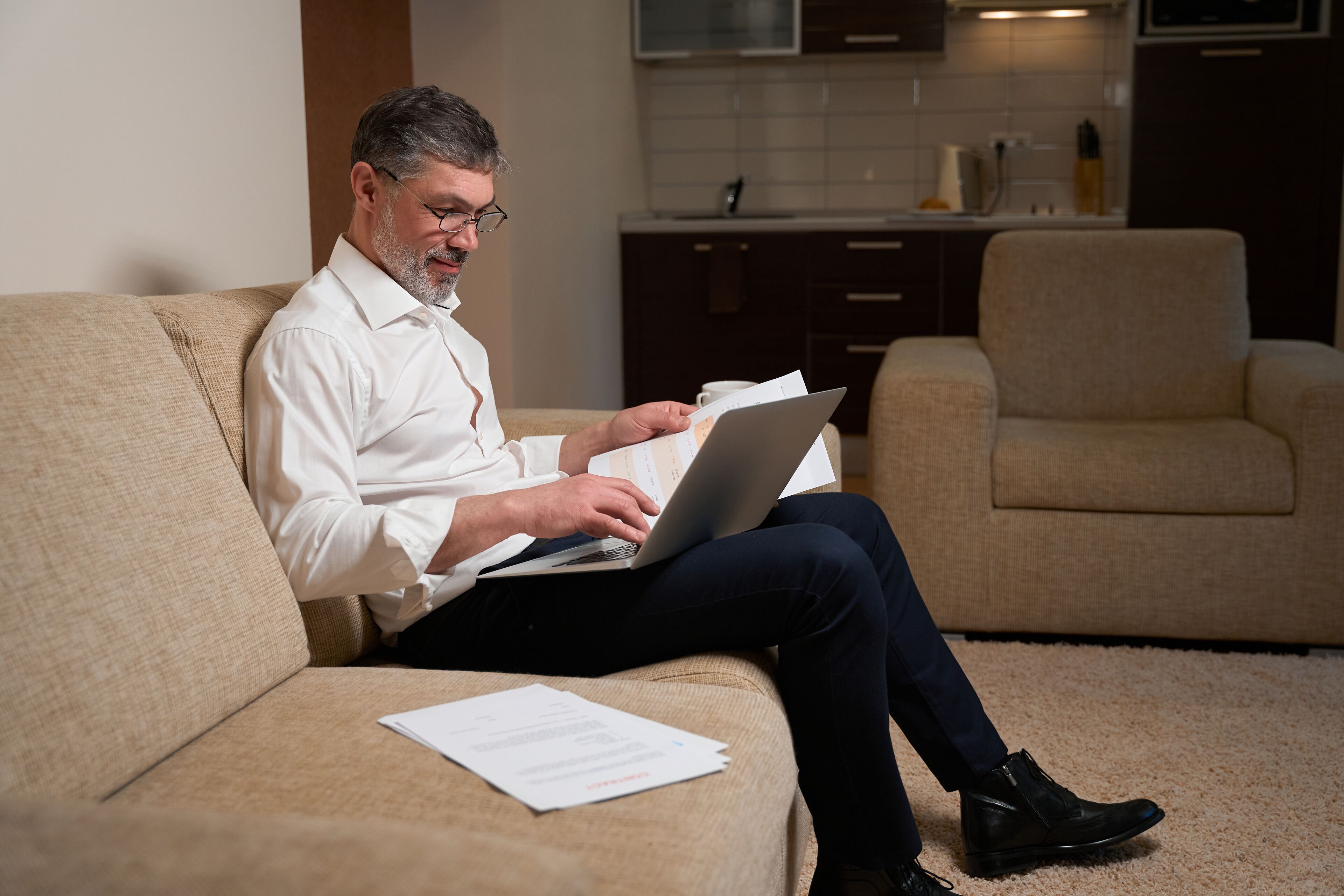 A man sitting on a beige sofa in a modern living room with a laptop and papers.