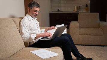 A man sitting on a beige sofa in a modern living room with a laptop and papers.