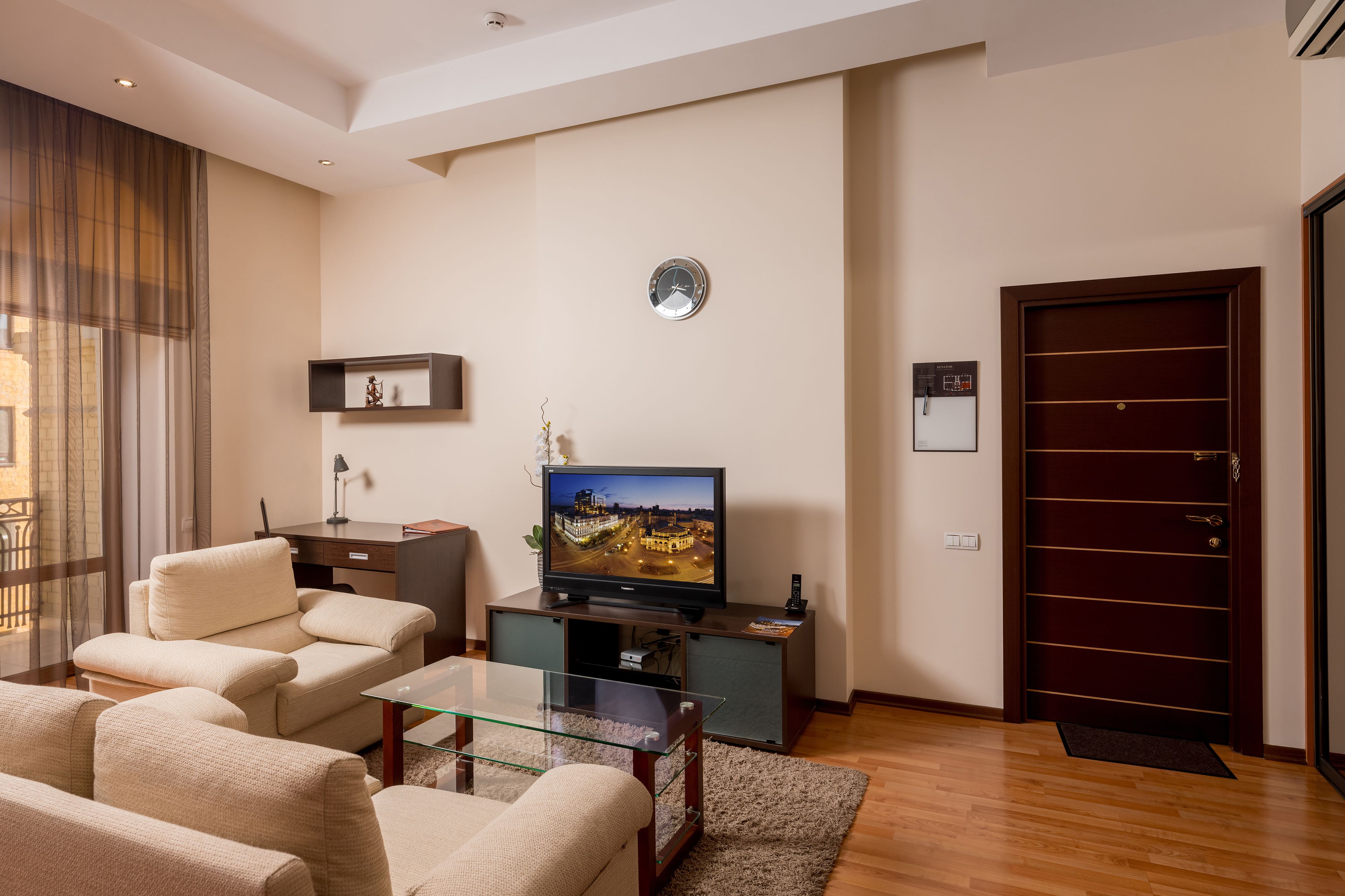 A modern living room with a flat-screen TV, beige sofas, a glass coffee table, and a dark wooden door.