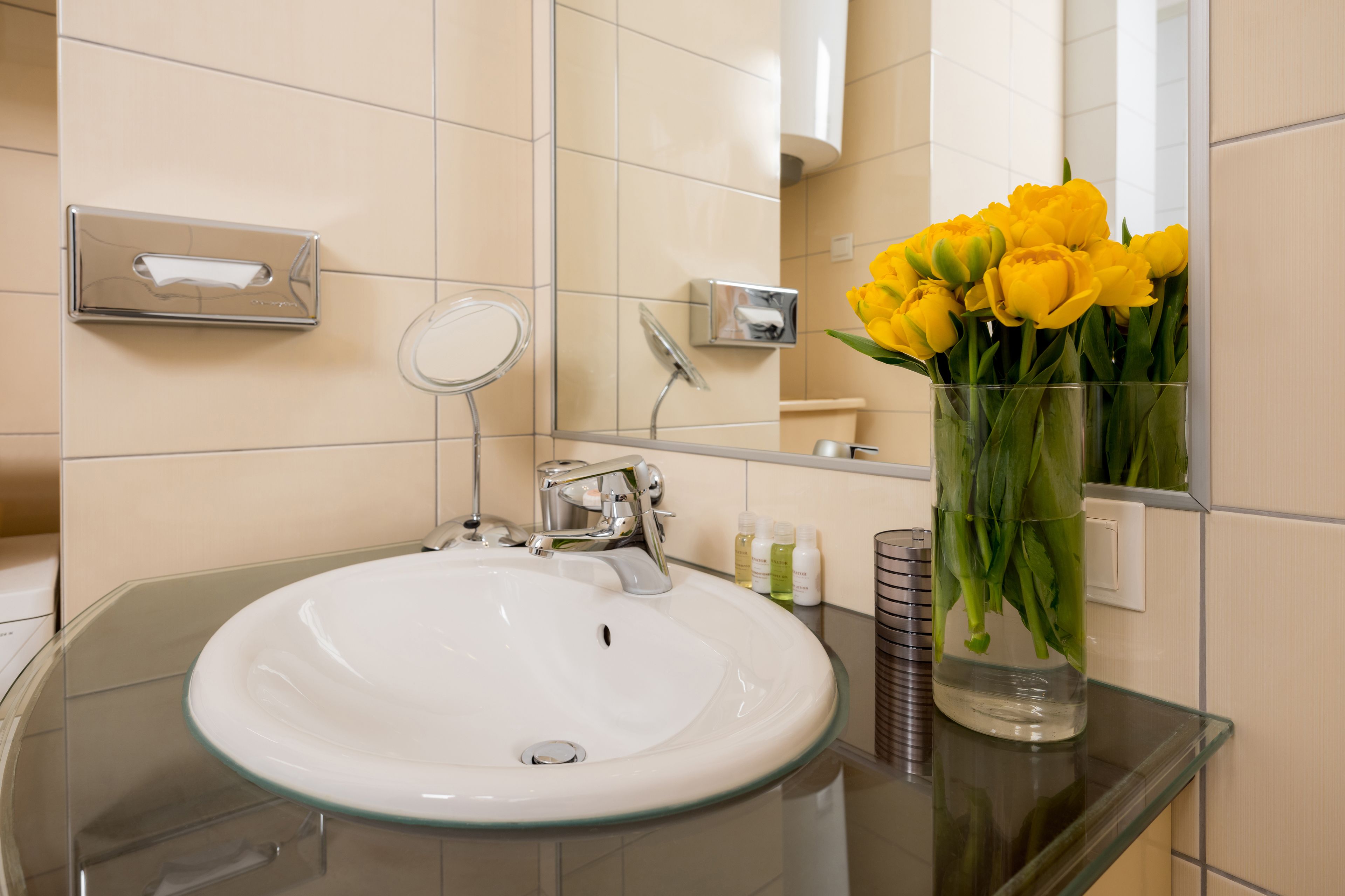 Ein Badezimmer mit einem Glaskonstruktion-Waschbecken, einem Spiegel und einer Vase mit gelben Blumen.