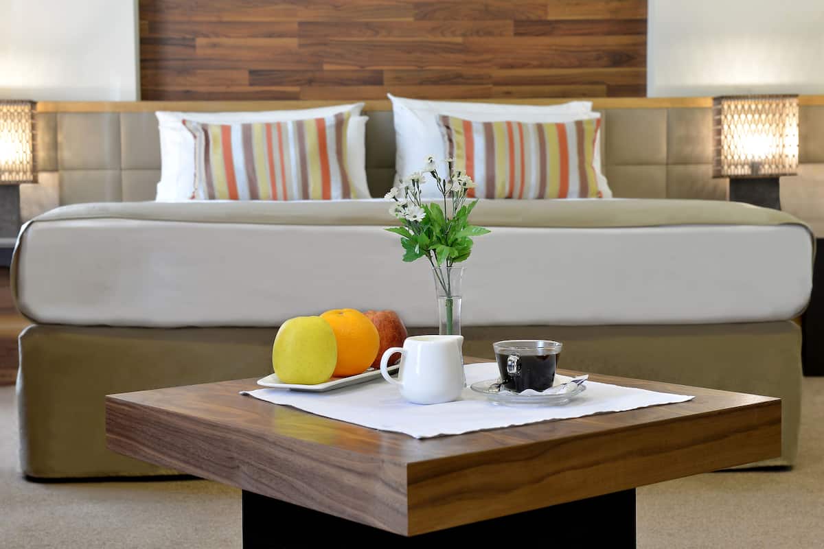 A neatly arranged breakfast table with fruits, a vase, and a cup in a hotel room with a bed and striped pillows in the background.