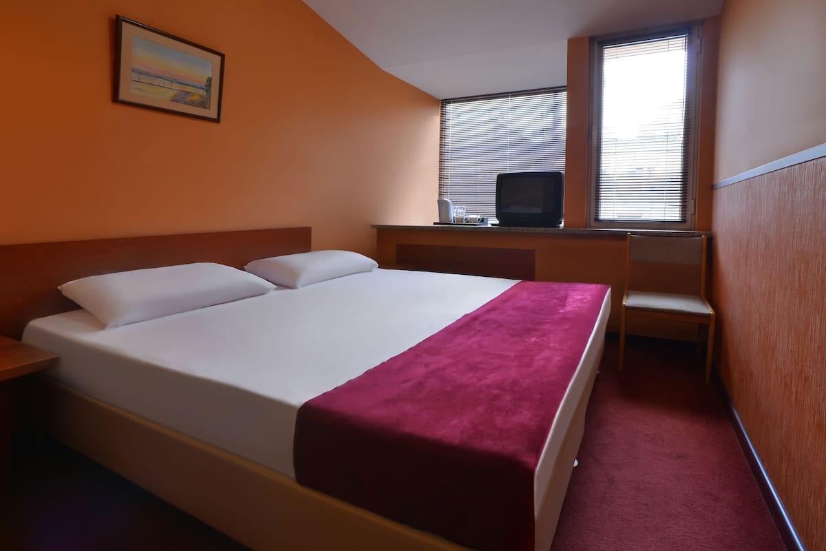 A hotel room with a bed, a desk with a television, and a window with blinds.