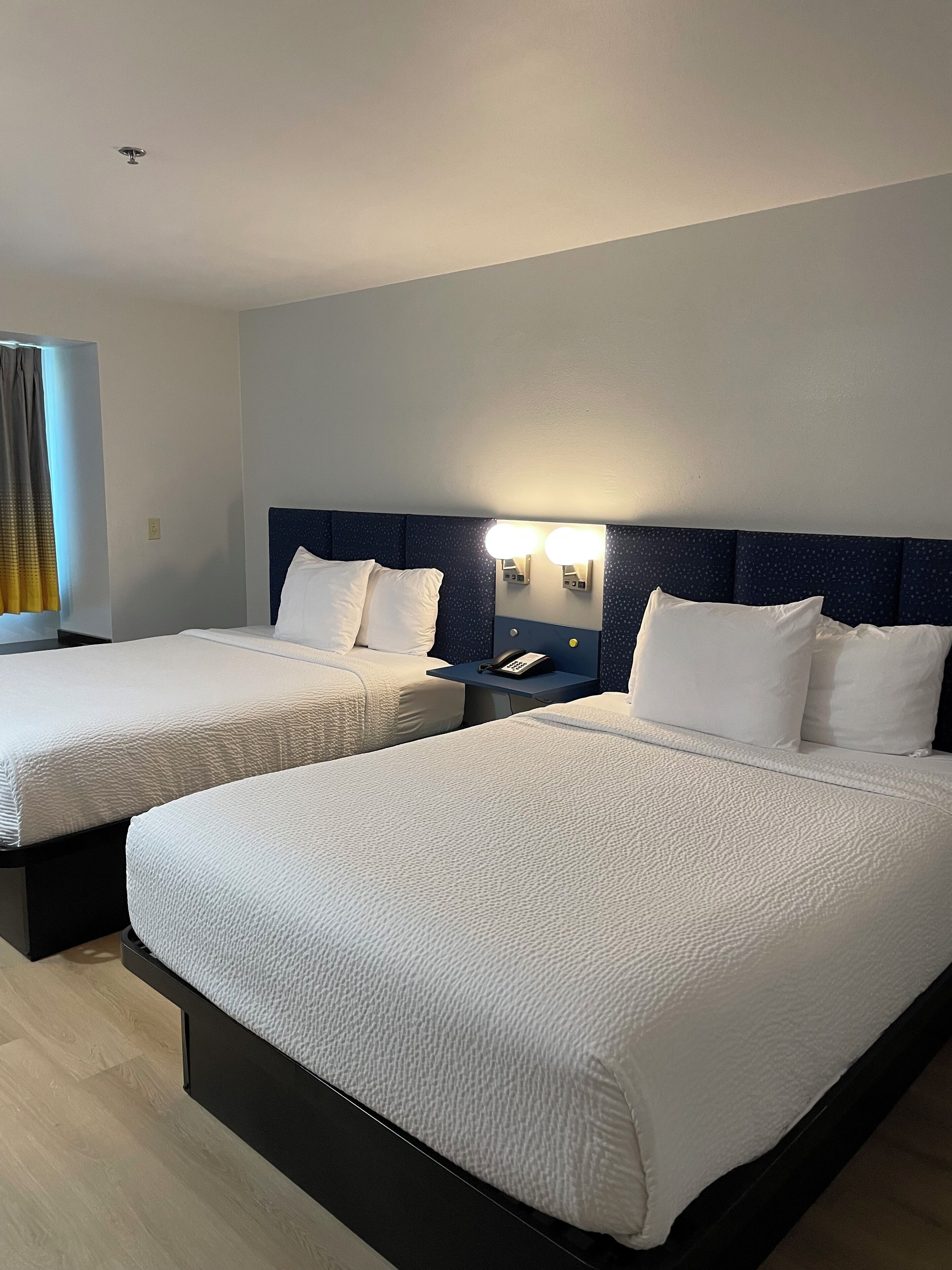 A hotel room with two beds, a nightstand with a phone, and a wall-mounted light fixture.