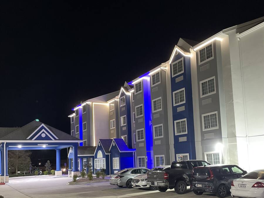 A multi-story hotel with a blue and white facade, a covered entrance, and a parking lot with several cars.