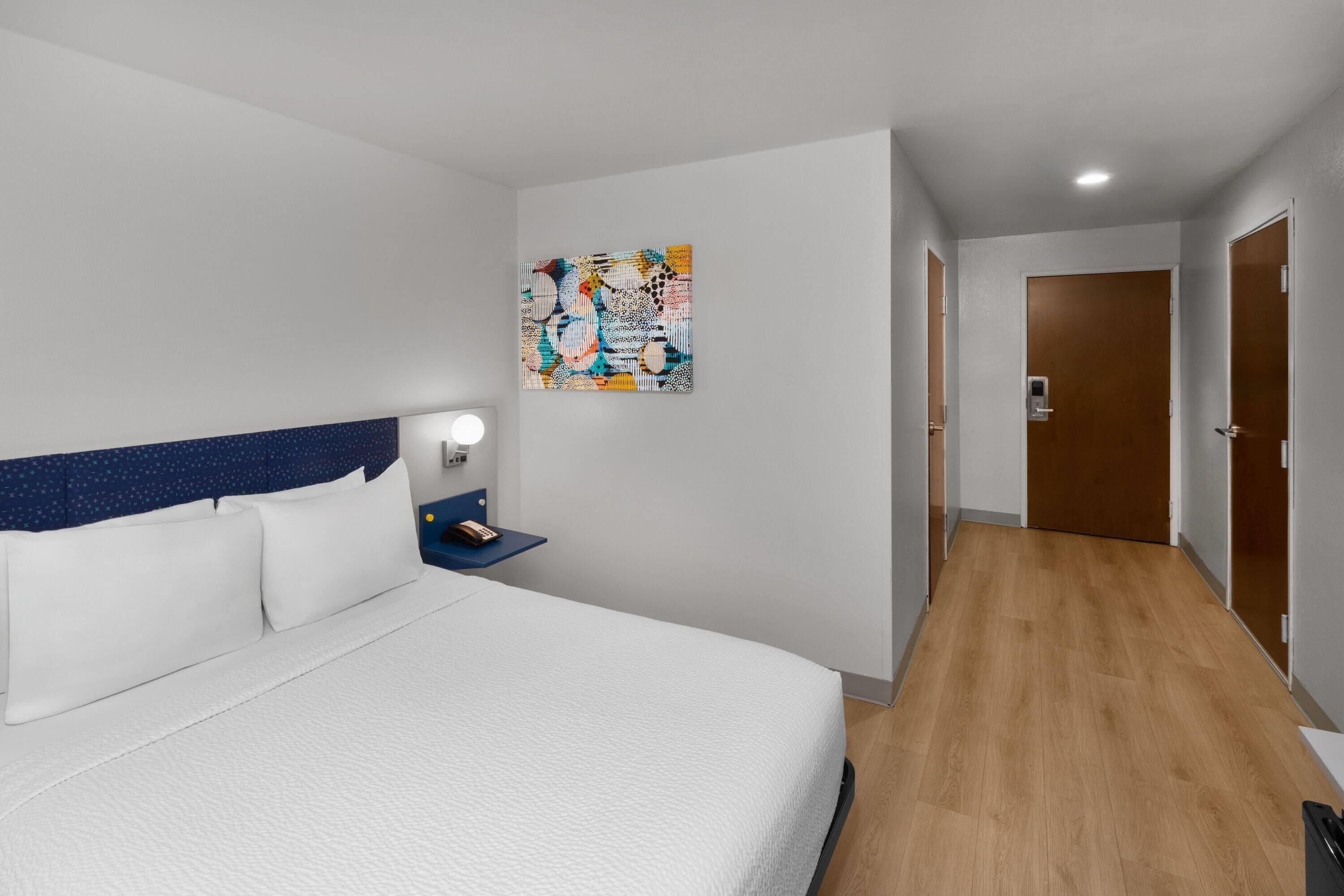 A hotel room with a bed, a nightstand, a wall art piece, and a wooden floor.