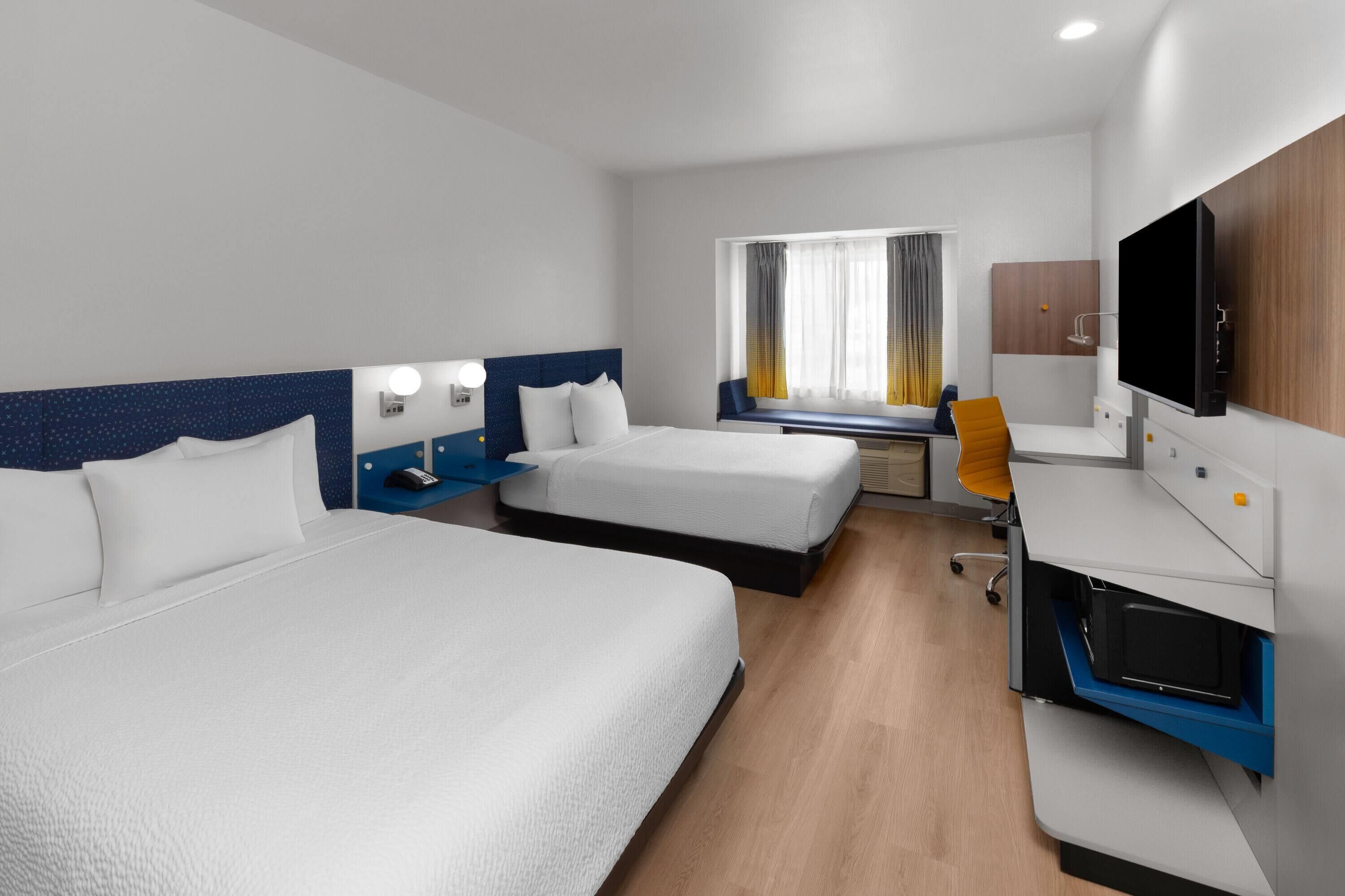 Ein Hotelzimmer mit zwei Betten, einem Schreibtisch, einem Fernseher und einem Fenster mit Vorhängen.
